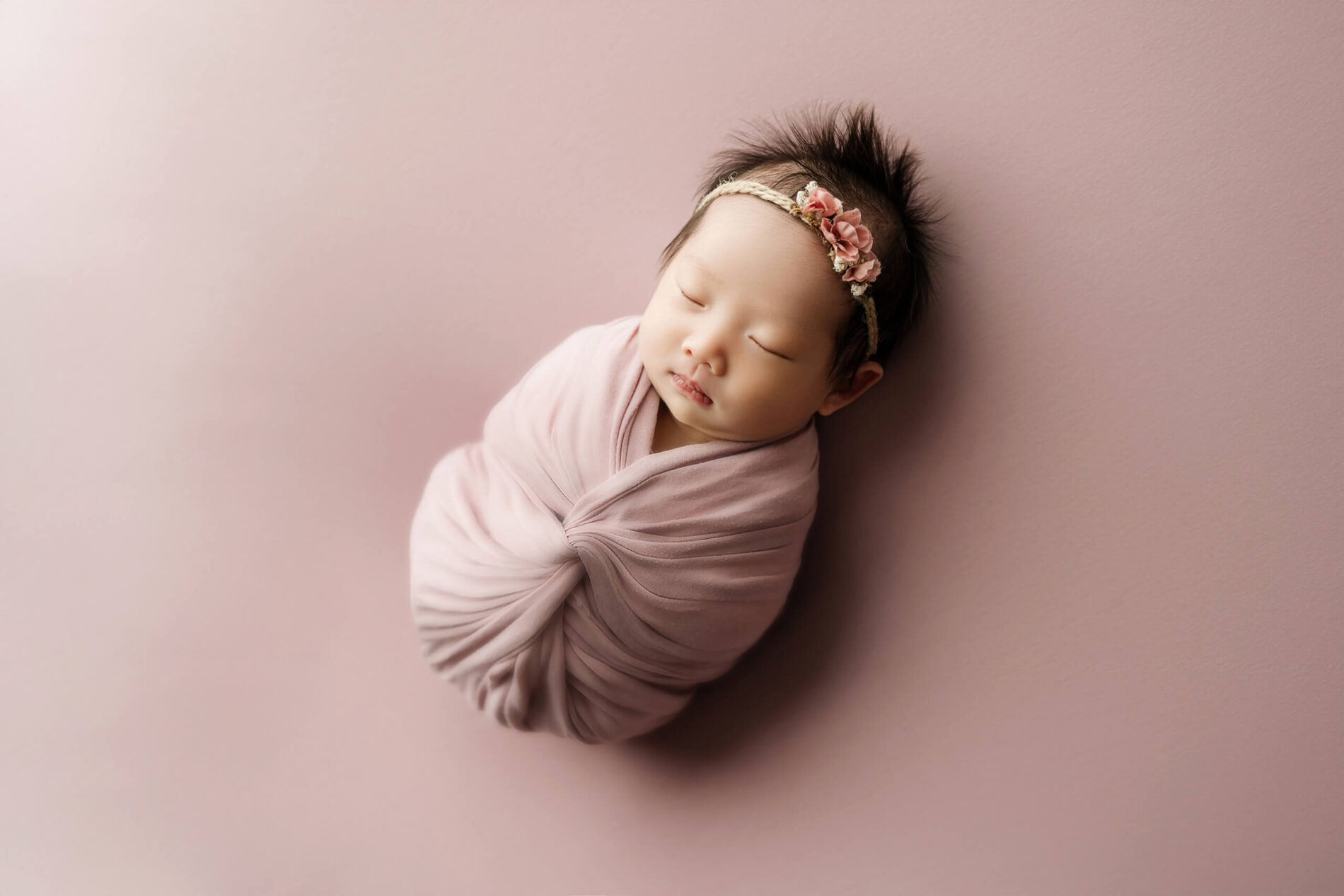Sleeping newborn wrapped in soft mauve pink with a floral headband against a matching pink backdrop.