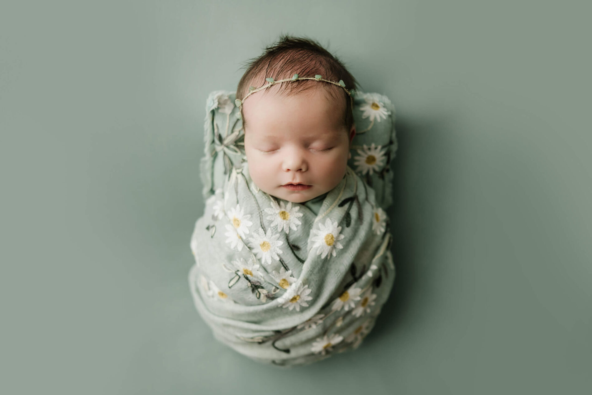 Sleeping newborn wrapped in sage green fabric with white daisies and wearing a delicate green leaf headband.