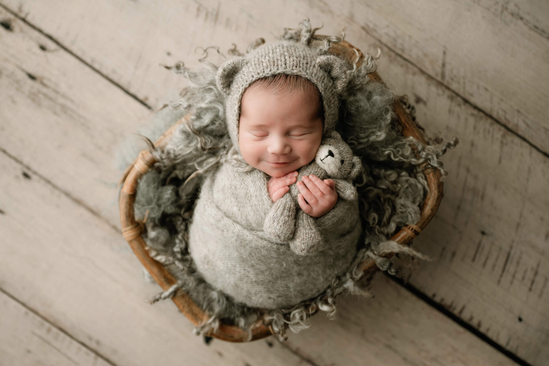 Smiling newborn wrapped in gray with a bear-ear bonnet and matching teddy bear, posed in a round basket with gray wool.