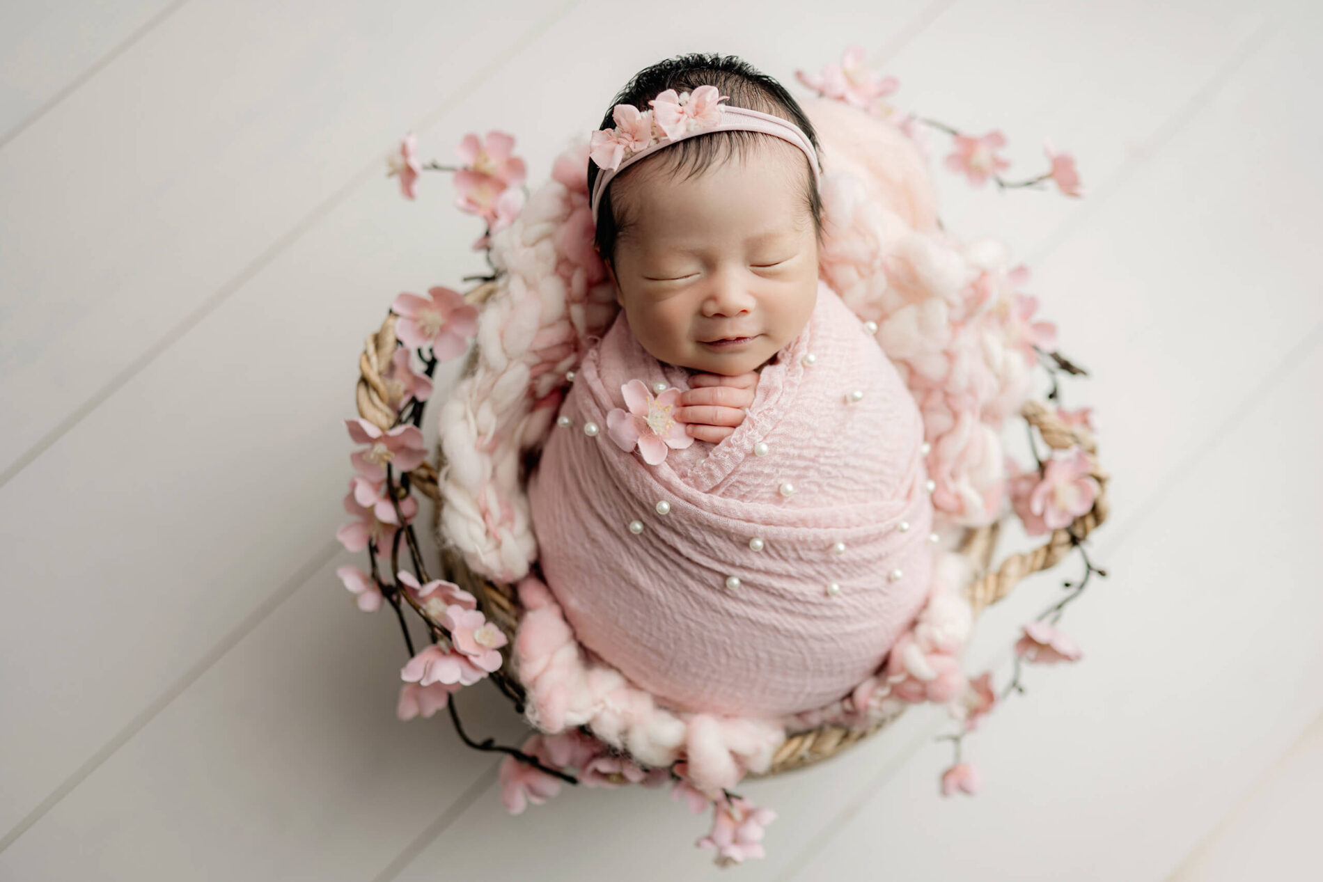 Sleeping newborn wrapped in blush pink with pearl accents and a floral headband, posed in a round basket surrounded by pink blossoms.