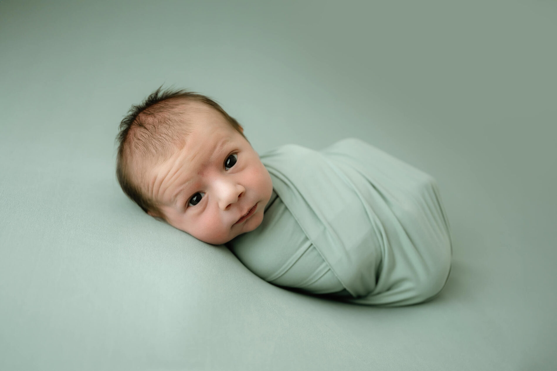 Awake newborn wrapped in soft mint green, lying on a matching studio backdrop and looking toward the camera.