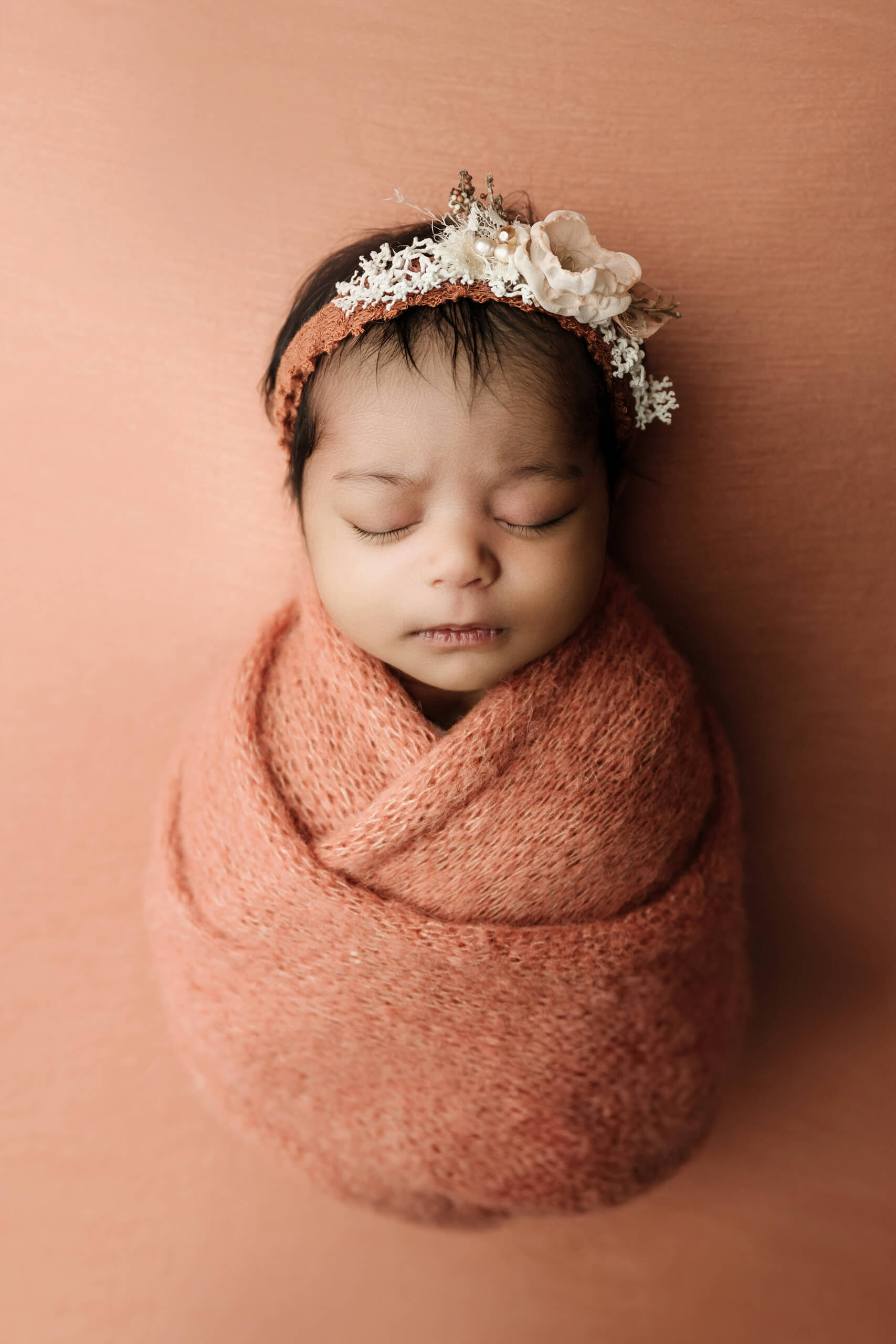 Sleeping newborn wrapped in rust-colored knit fabric with a floral headband against a matching warm peach backdrop.