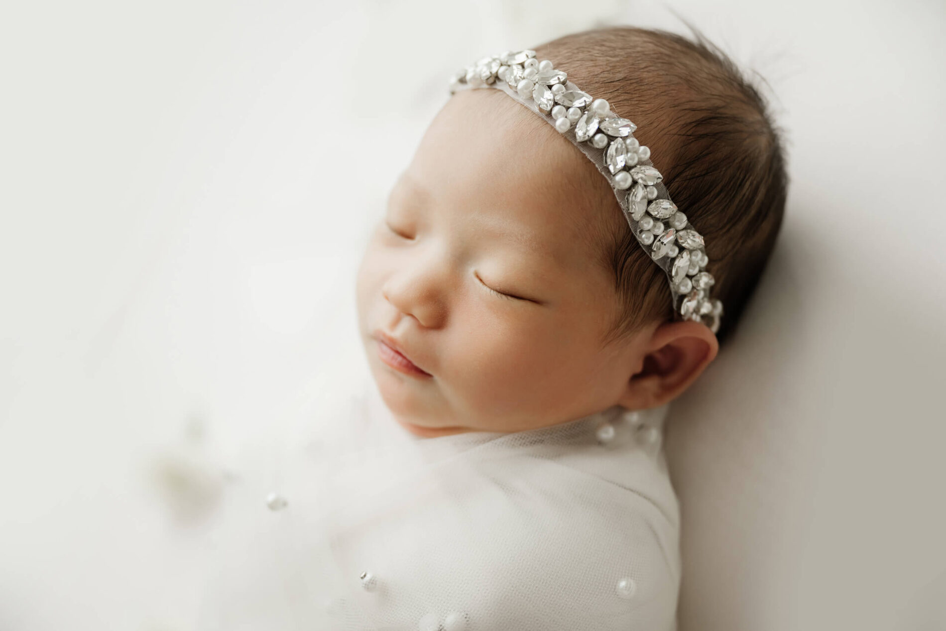Close-up of a sleeping newborn wrapped in white and wearing a jeweled pearl headband on a soft white backdrop.