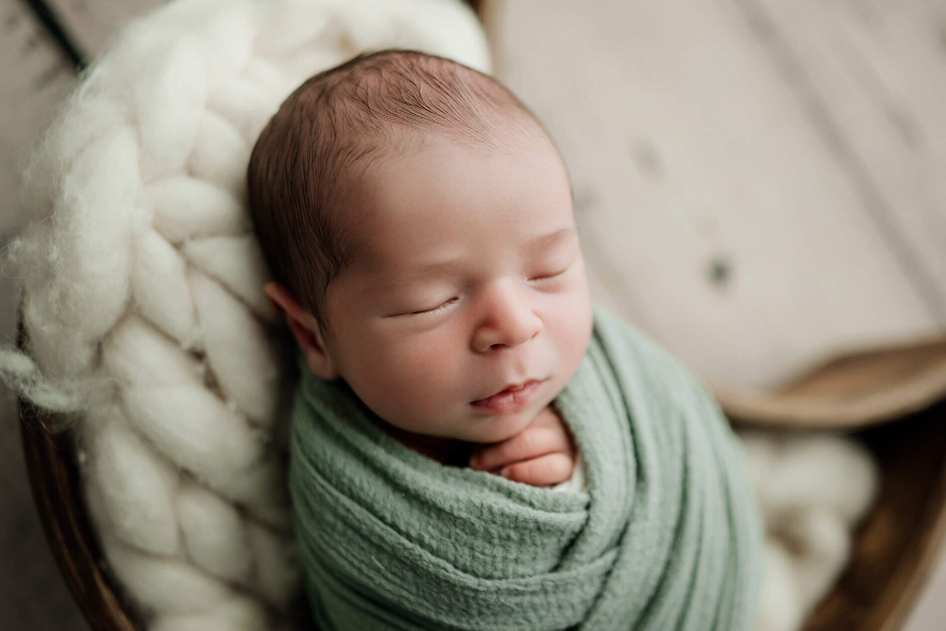 Sleeping newborn wrapped in sage green, posed in a wooden bowl lined with soft cream wool.