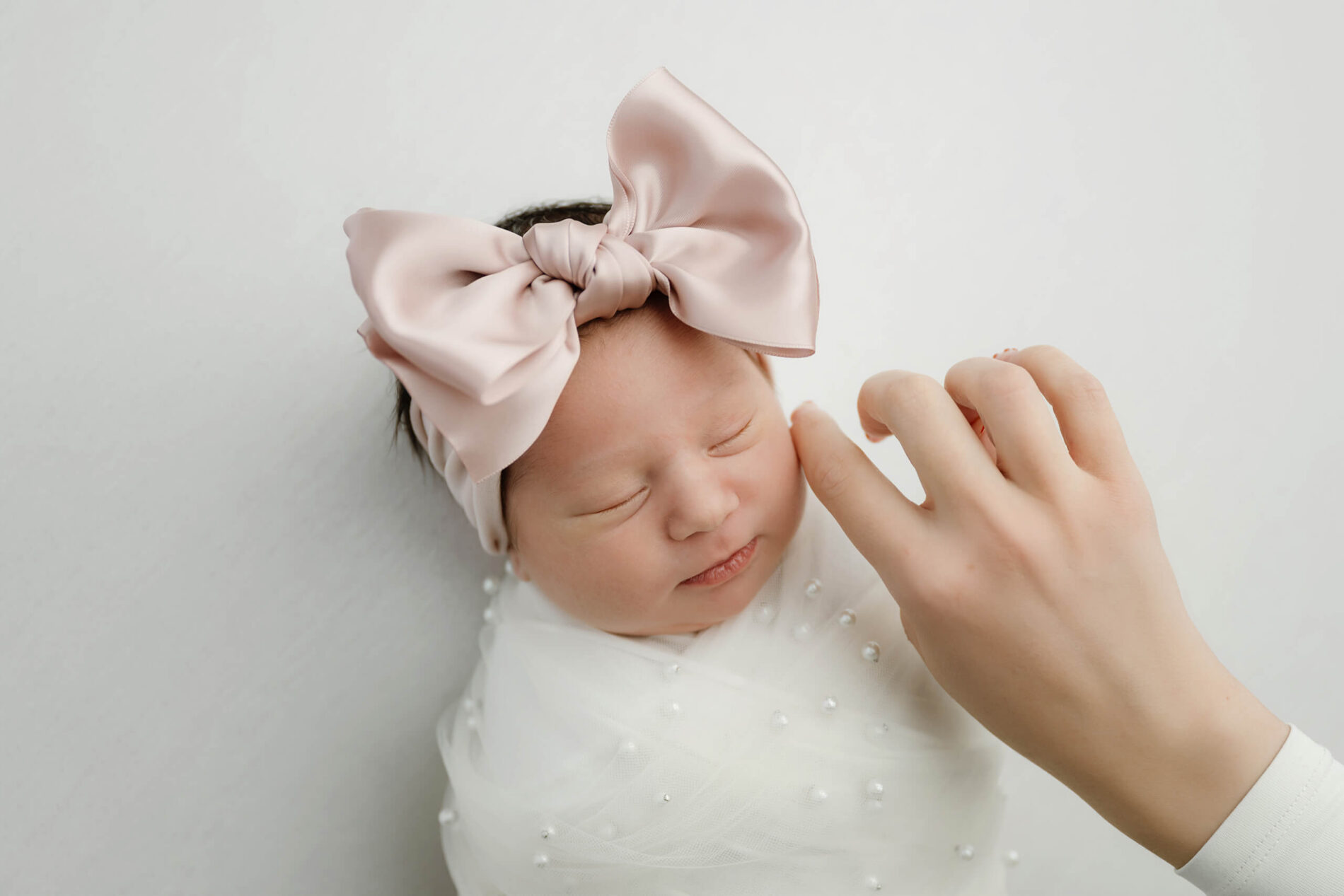 Sleeping newborn wrapped in white with pearl accents and an oversized blush bow as an adult gently touches the baby’s cheek.
