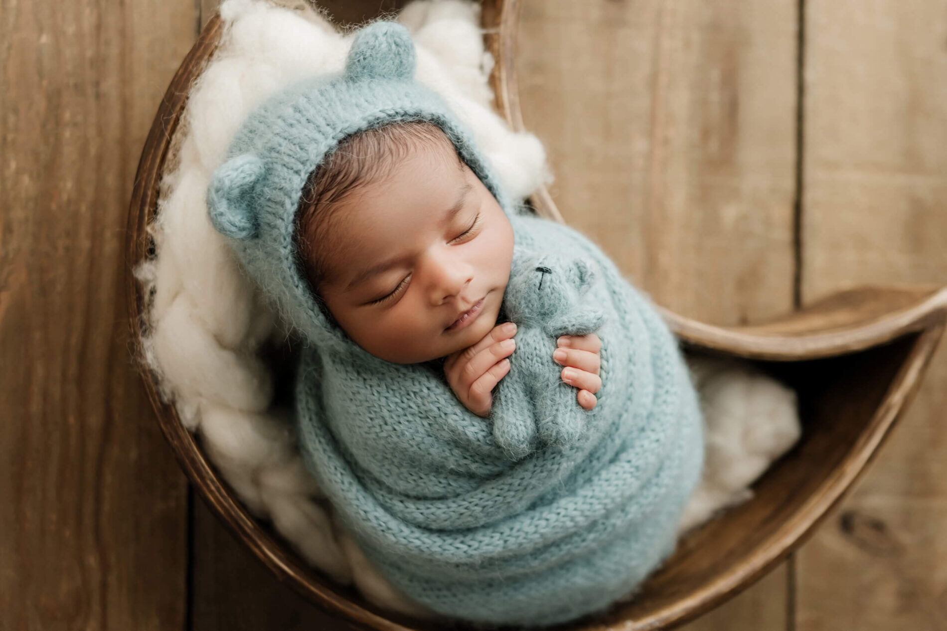 Sleeping newborn wrapped in powder blue with a bear-ear bonnet and matching teddy bear, posed in a wooden crescent moon prop.
