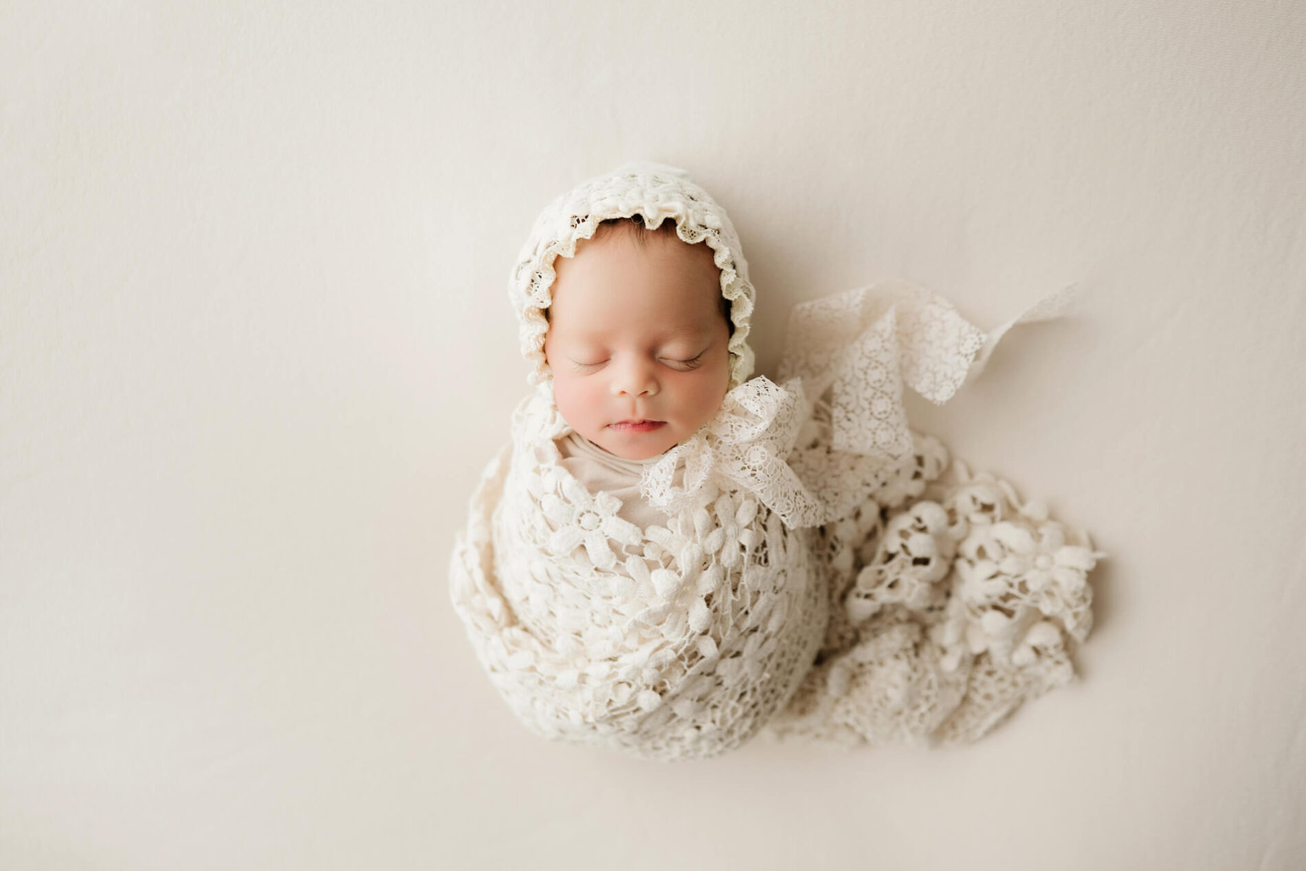 Sleeping newborn wrapped in cream lace with a matching lace bonnet and trailing ribbons on a neutral studio backdrop.
