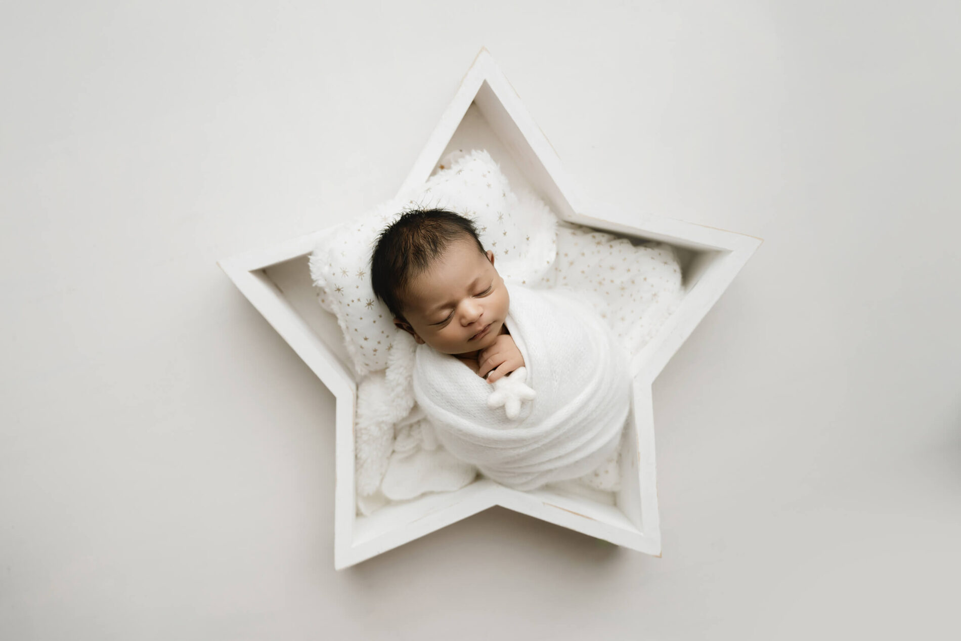 Sleeping newborn wrapped in white and posed inside a white wooden star with star-patterned bedding and a small plush star.