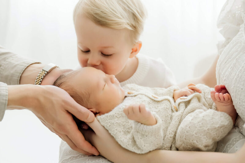 Older brother gently kisses a newborn baby held in a parent’s arms during a bright studio session.