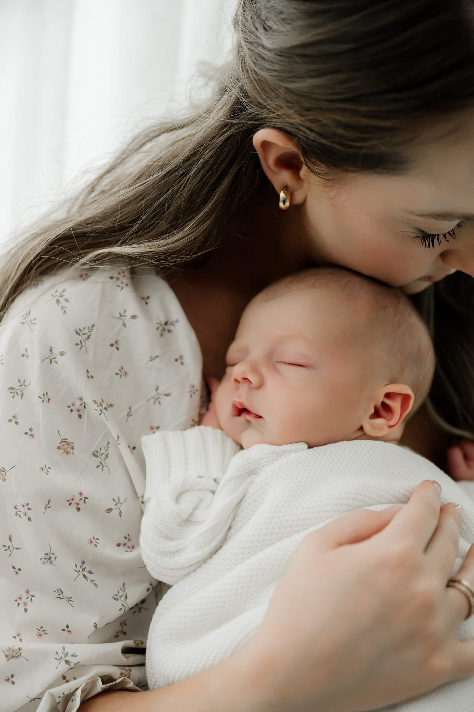 Mother kisses her sleeping newborn’s head while holding the baby close during a softly lit studio session.