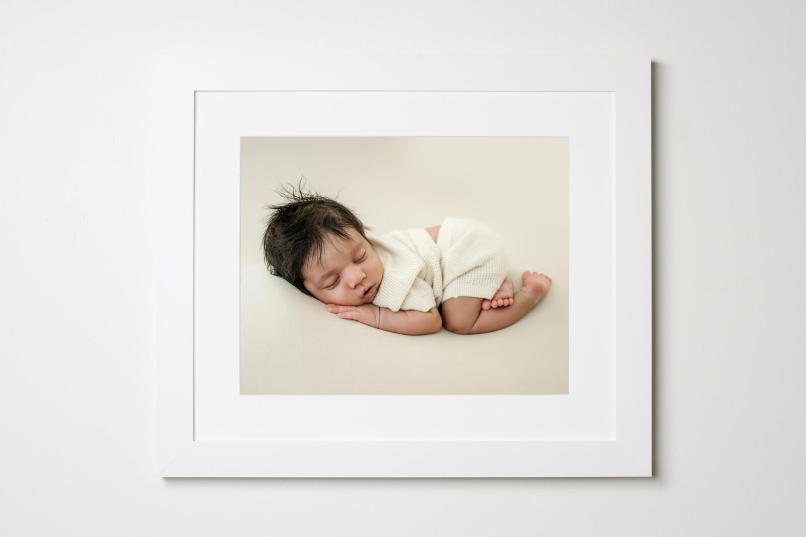 White-framed wall art featuring a sleeping newborn in a cream outfit, displayed against a light neutral wall.