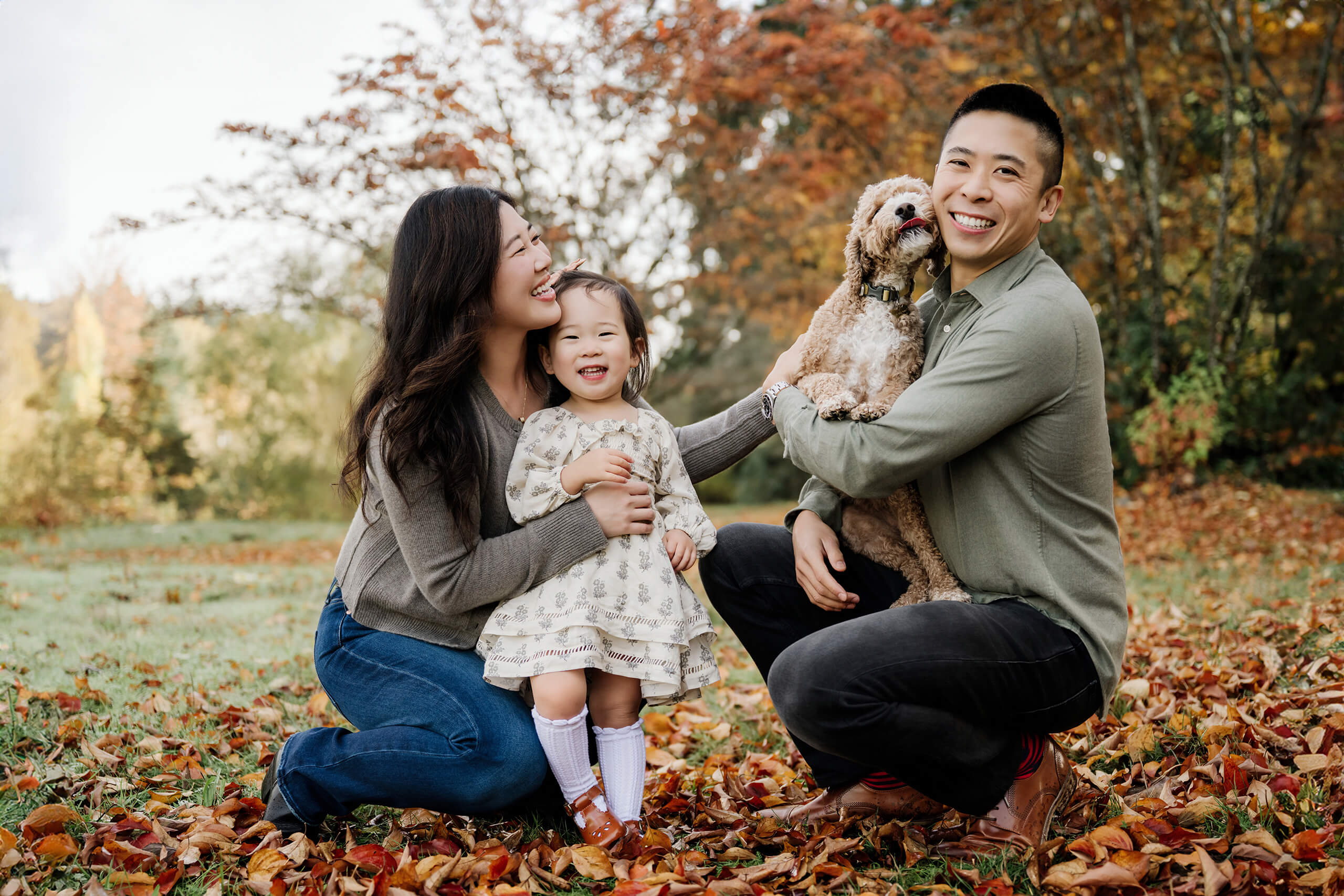 Family with young daughter and dog poses together among red and orange fallen leaves.