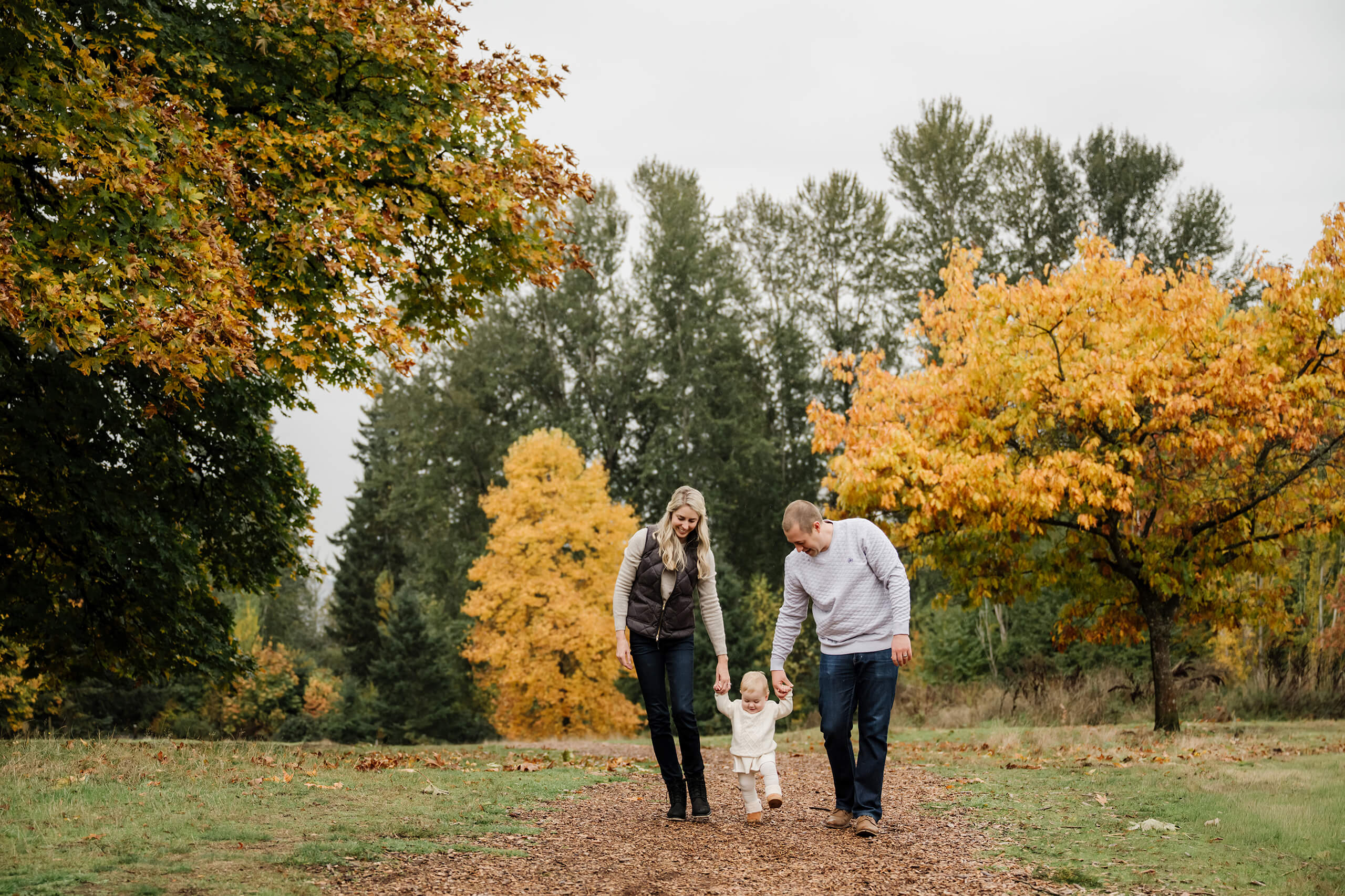 Parents hold their baby&rsquo;s hands while walking along a path framed by colorful fall trees.