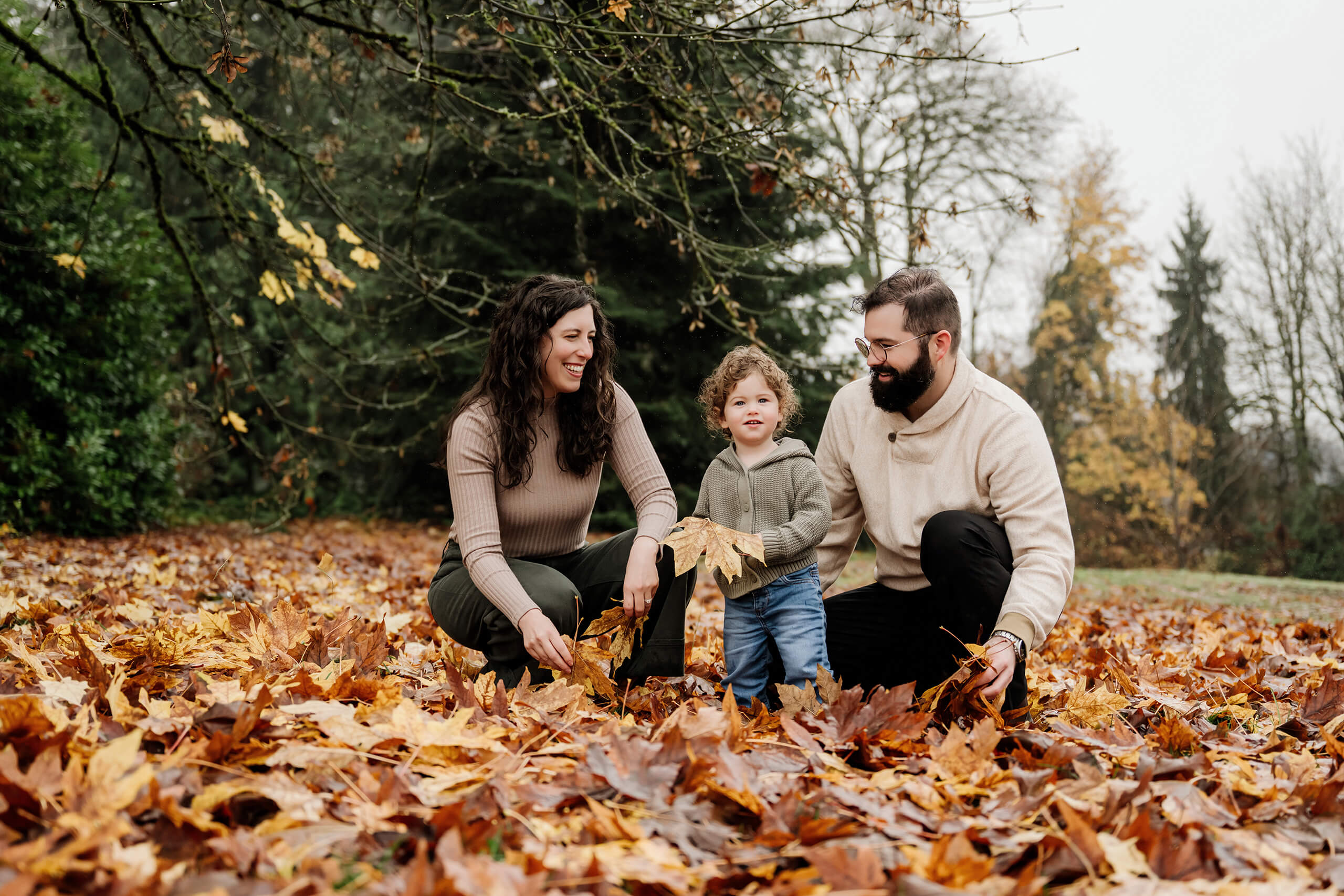 Parents crouch beside their young child playing with fallen leaves in an autumn park setting.
