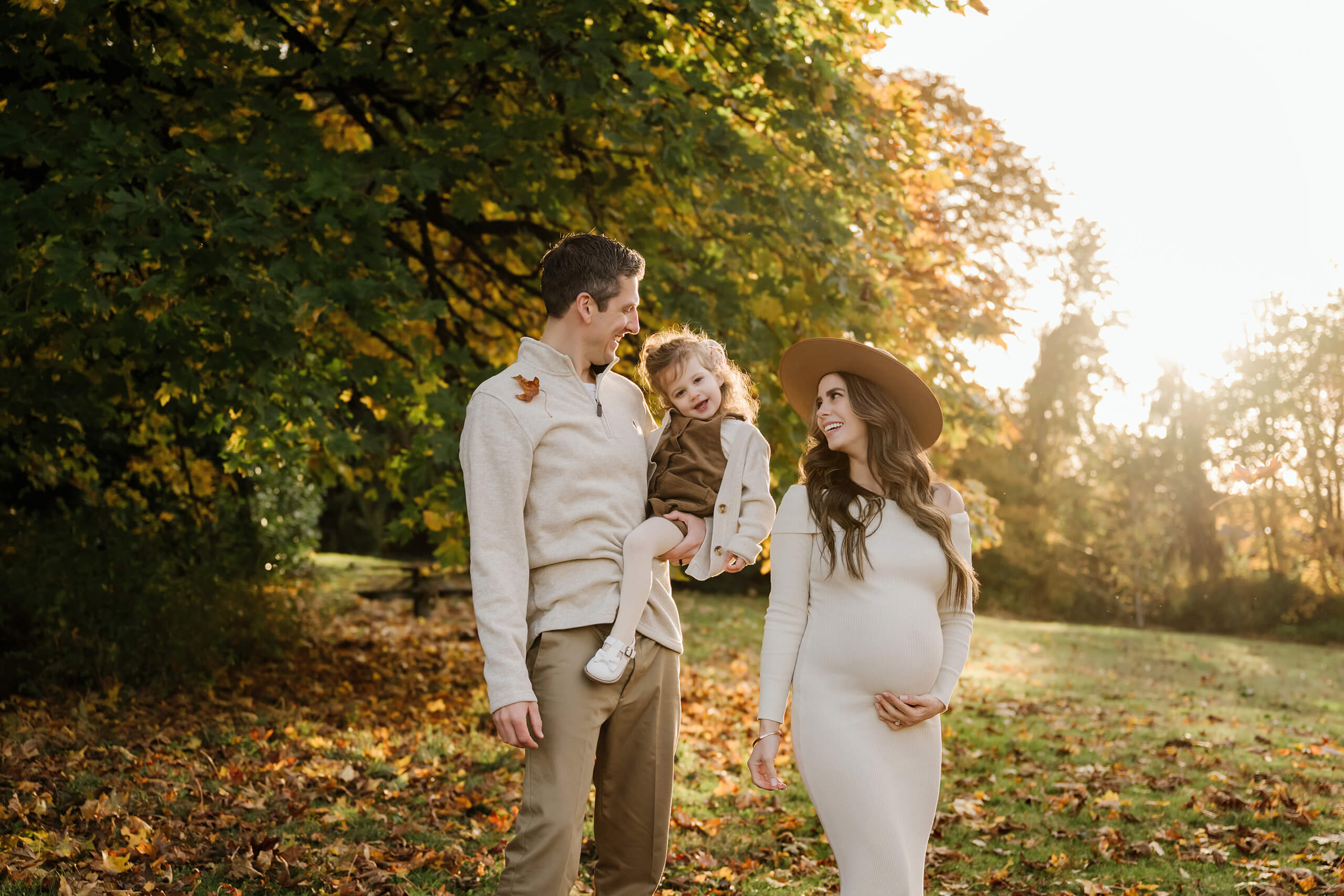 Expecting mom walks beside her partner carrying their daughter in warm evening light and fall foliage.