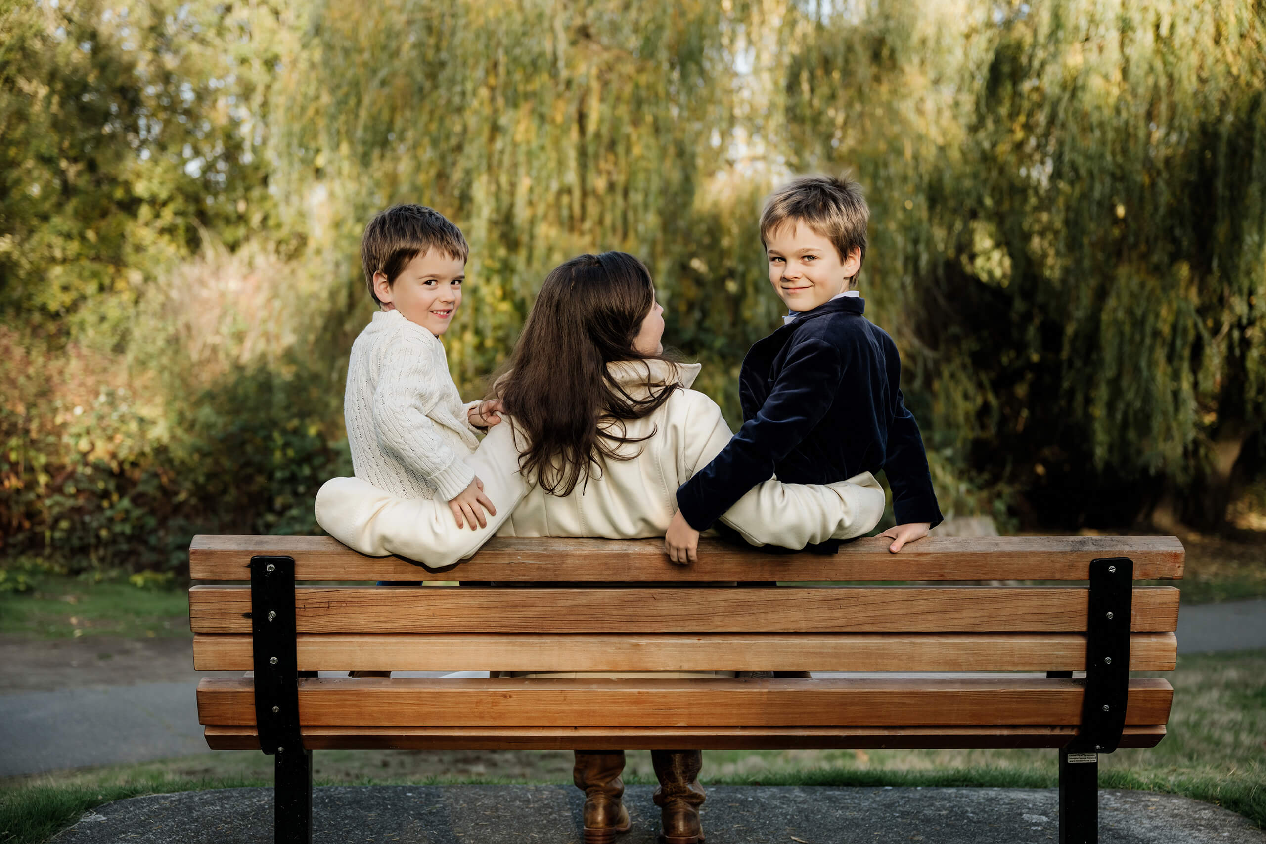 Mom sits between her two sons on a park bench surrounded by early fall greenery.