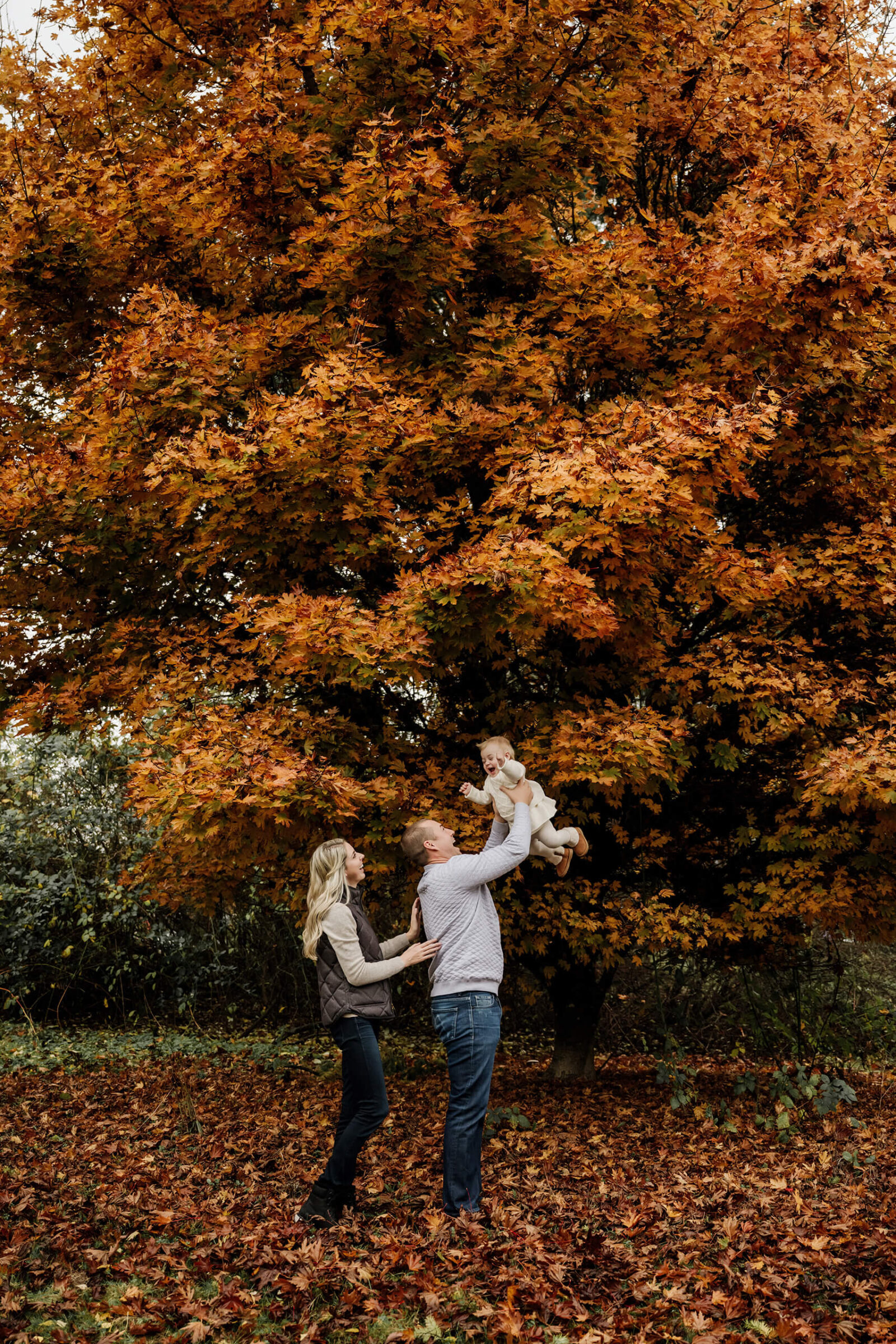 Dad lifts his baby overhead beside mom beneath a large tree filled with orange autumn leaves.