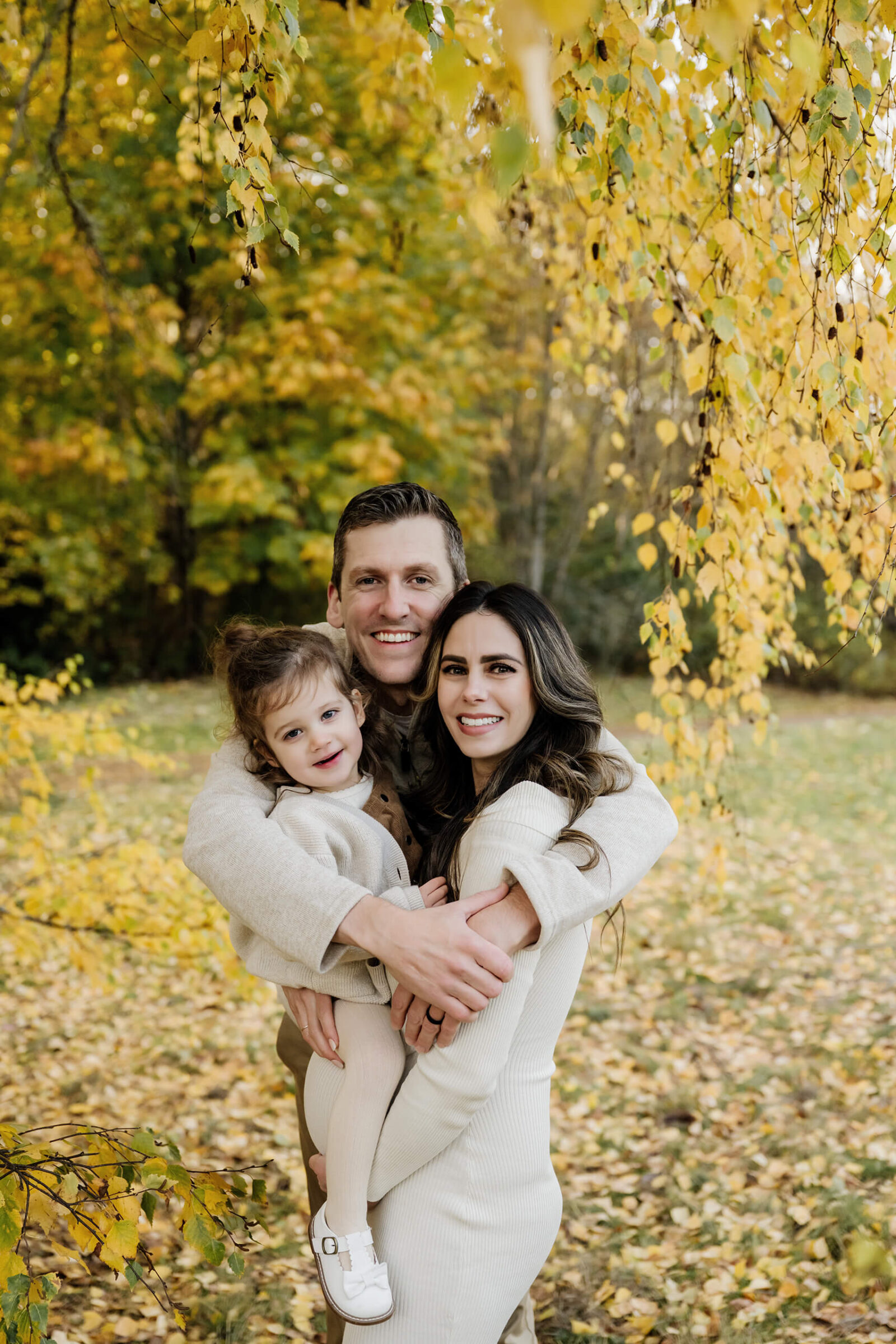 Parents cuddle their young daughter beneath hanging golden leaves during a fall family session.