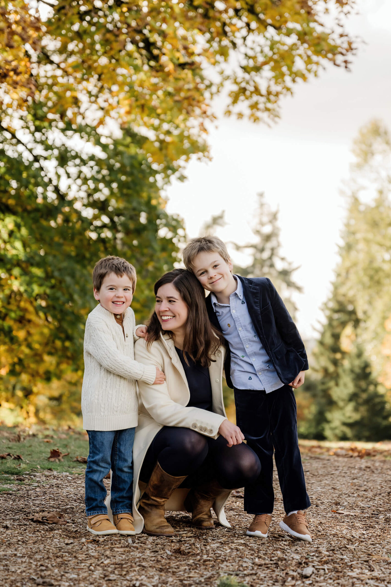 Mom crouches between her two sons beneath trees with yellow and green fall foliage.