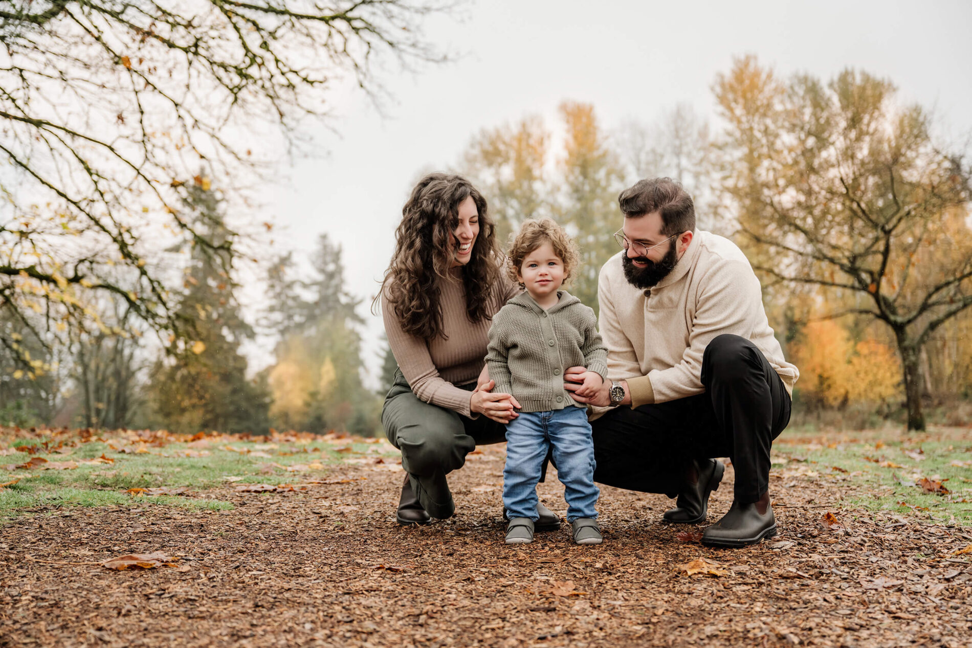 Parents crouch beside their young son on a park path surrounded by soft autumn color.