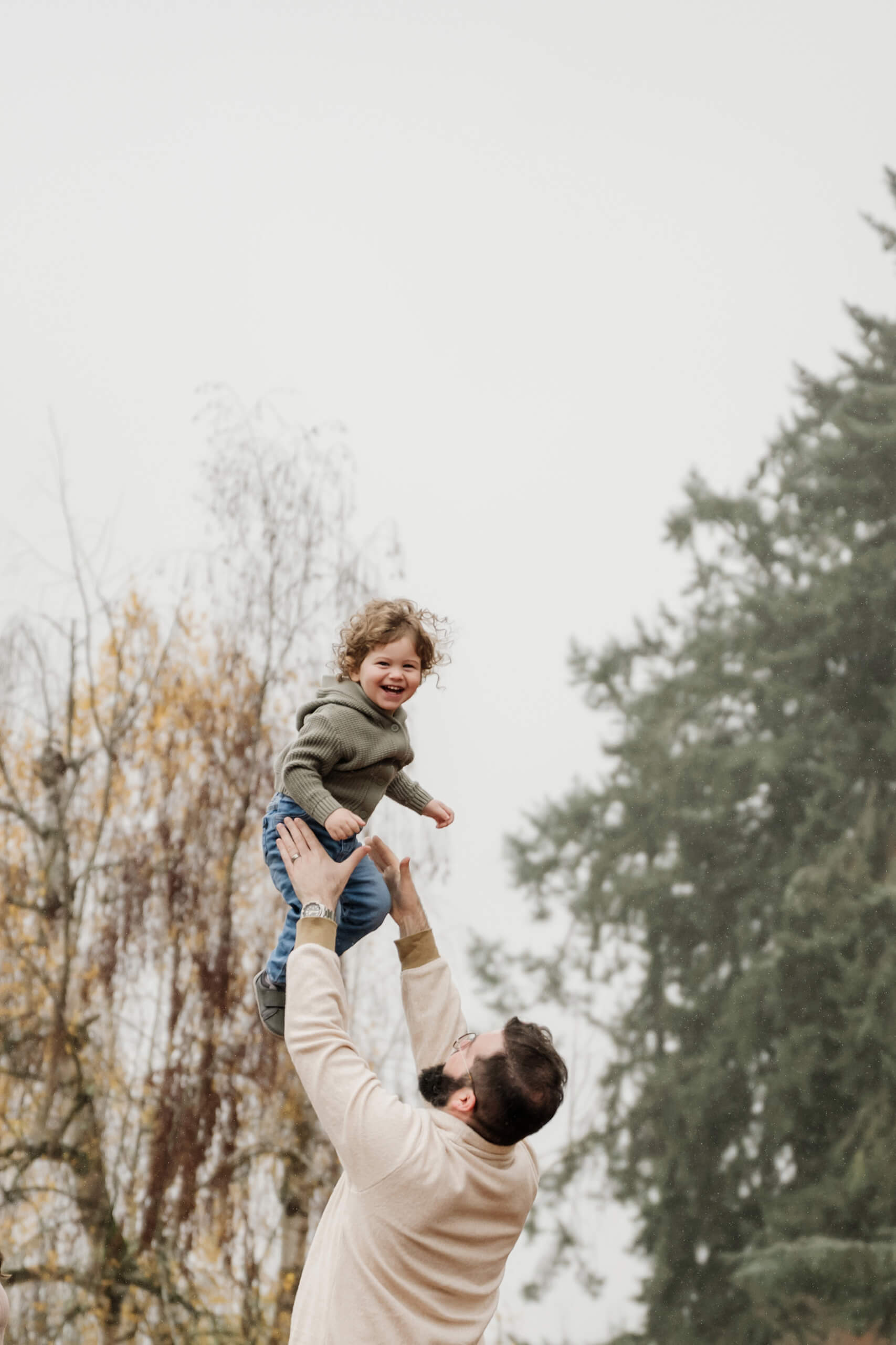 Dad lifts his laughing young son overhead against a backdrop of late-autumn trees.