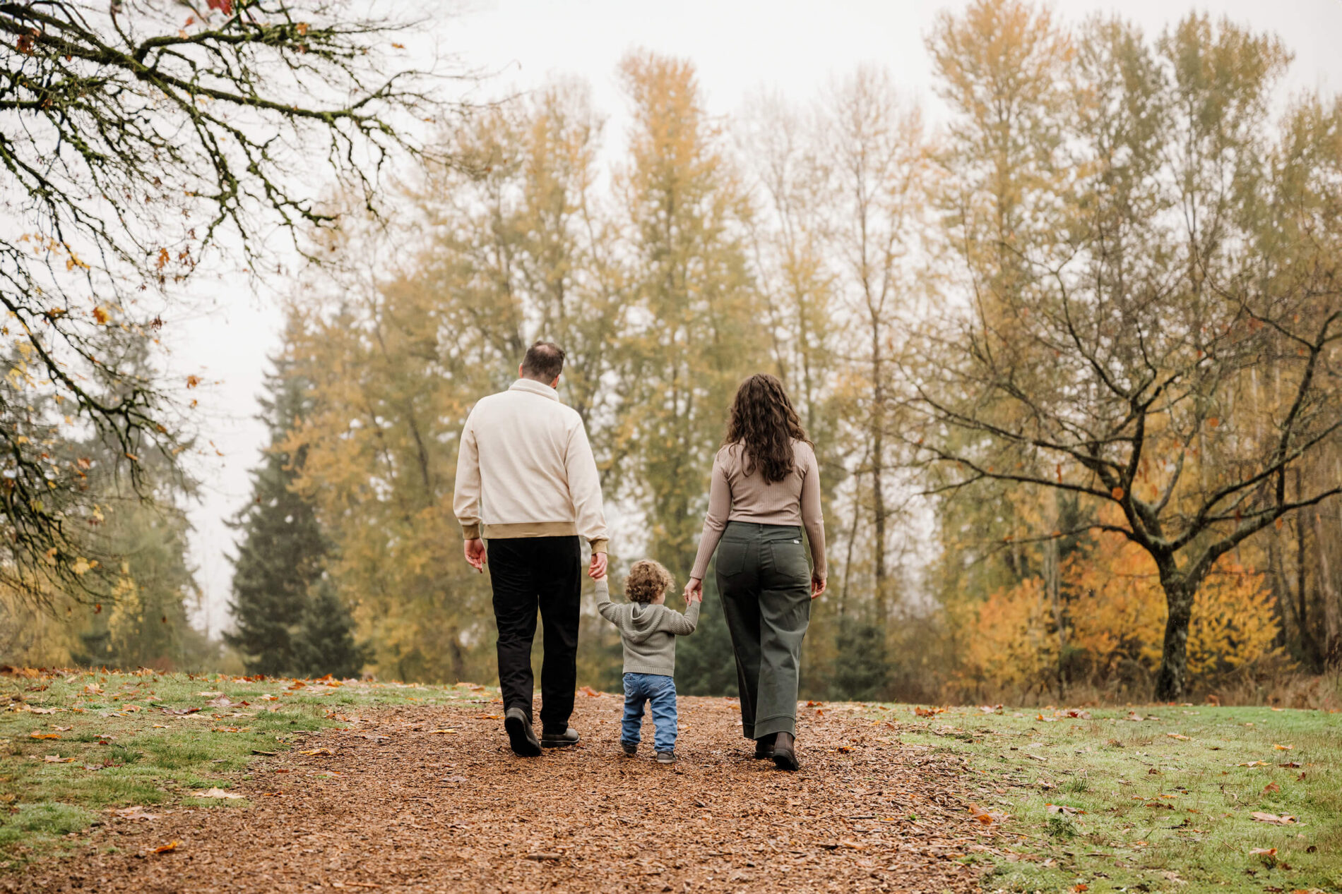 Parents walk away holding their young son’s hands along a path lined with fall trees.