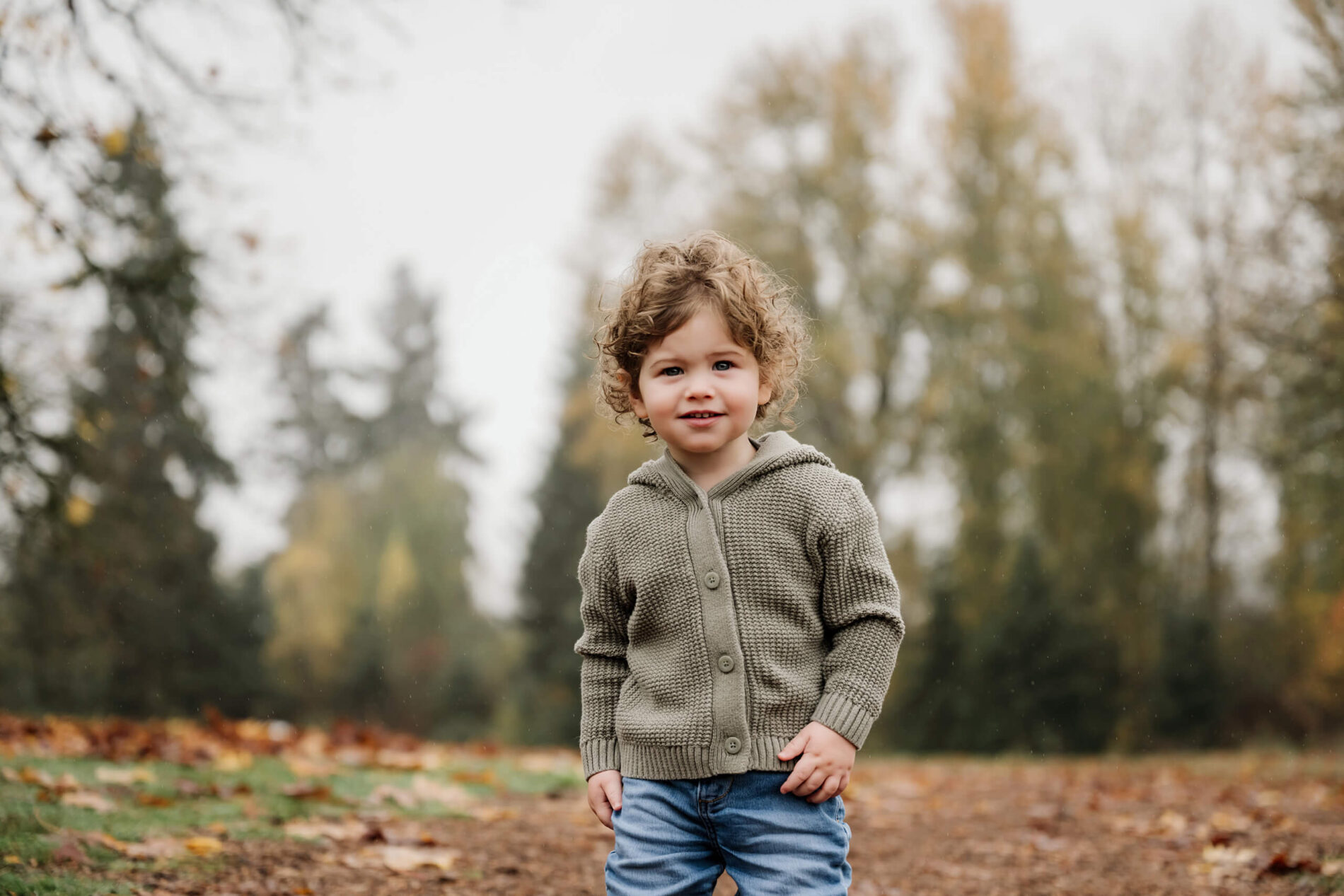 Young boy stands on a park path with fallen leaves and muted autumn trees behind him.