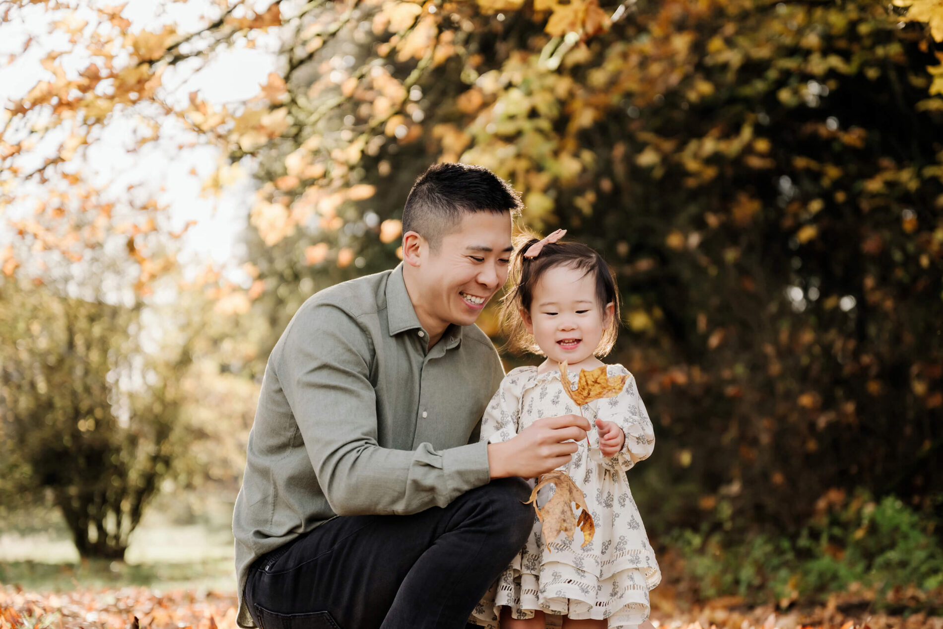 Dad crouches beside his daughter as she holds fallen autumn leaves.