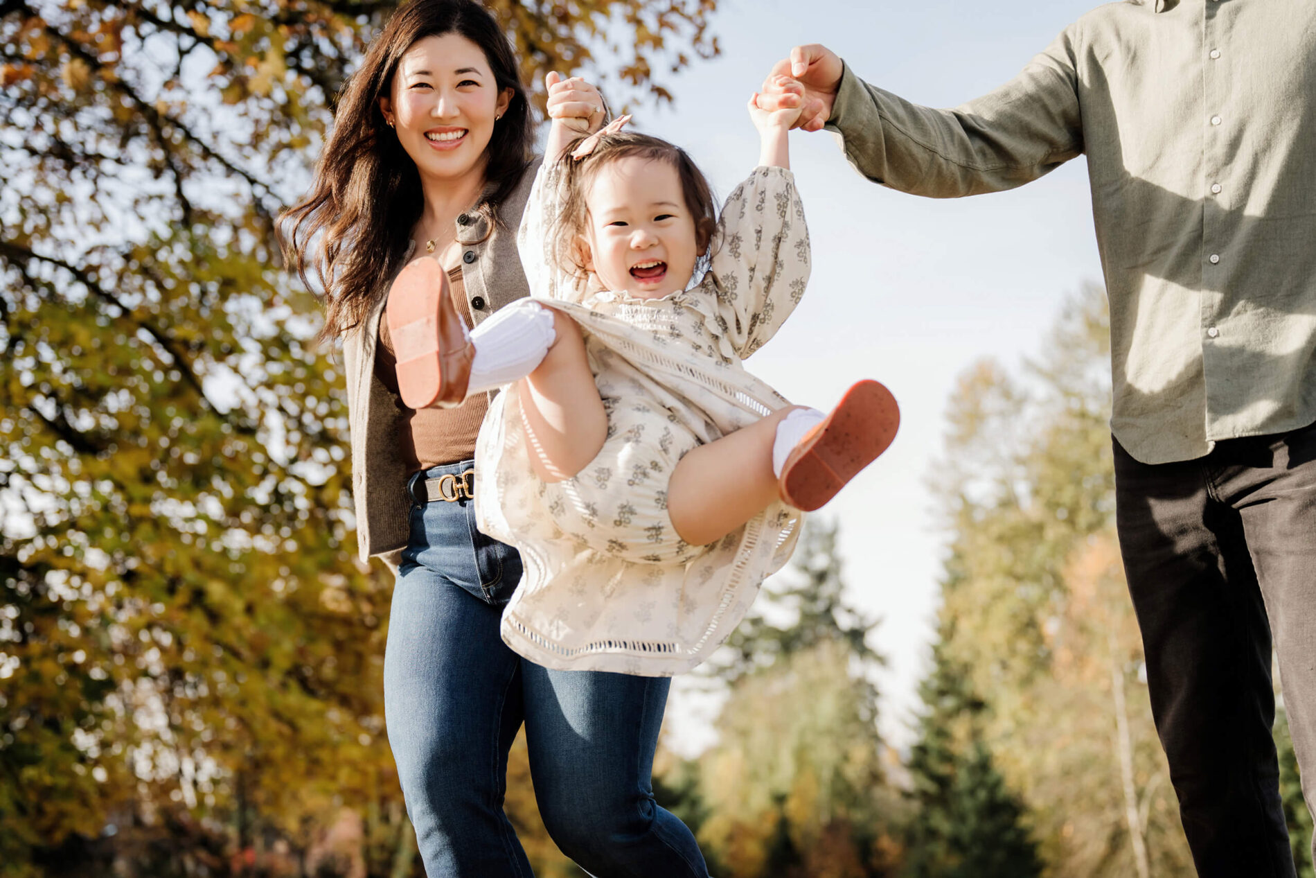 Parents swing their laughing daughter by the hands with fall foliage behind them.