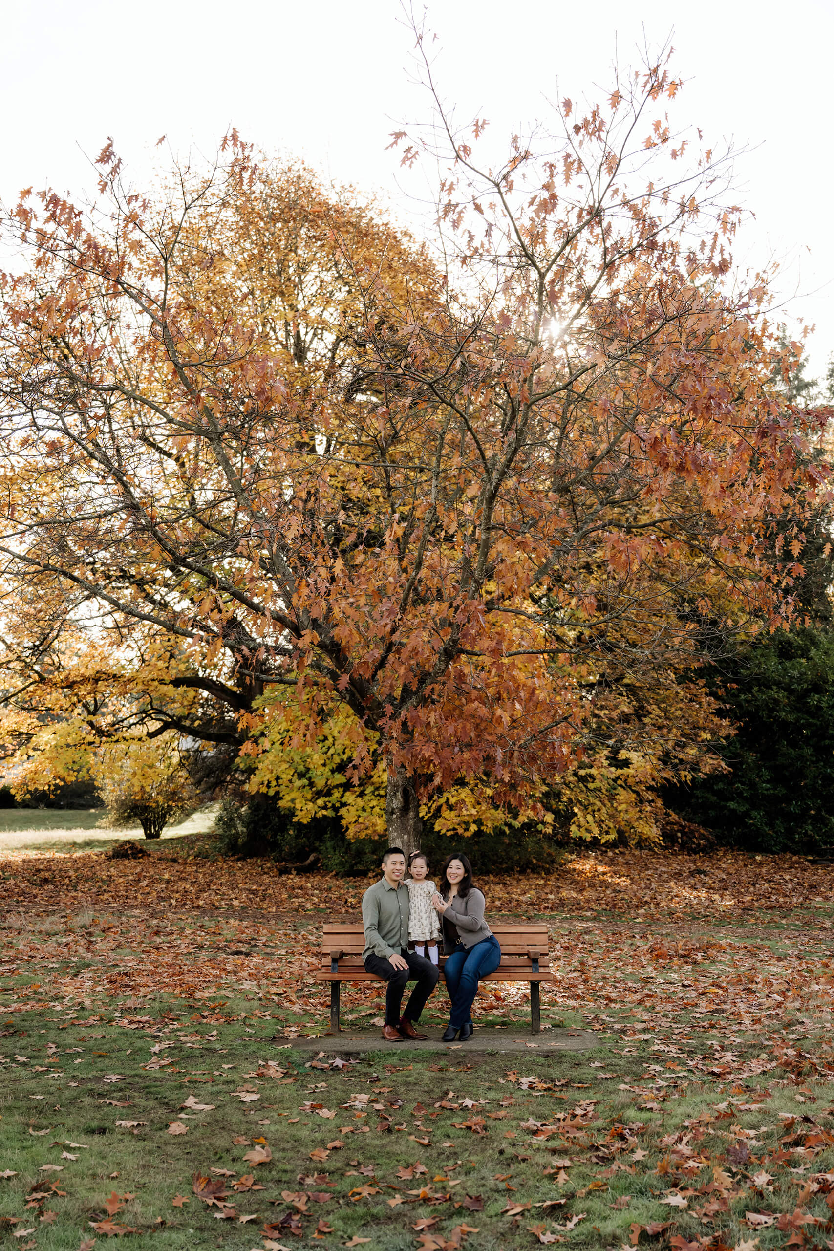 Family of three sits on a park bench beneath a large tree with orange and yellow autumn leaves.