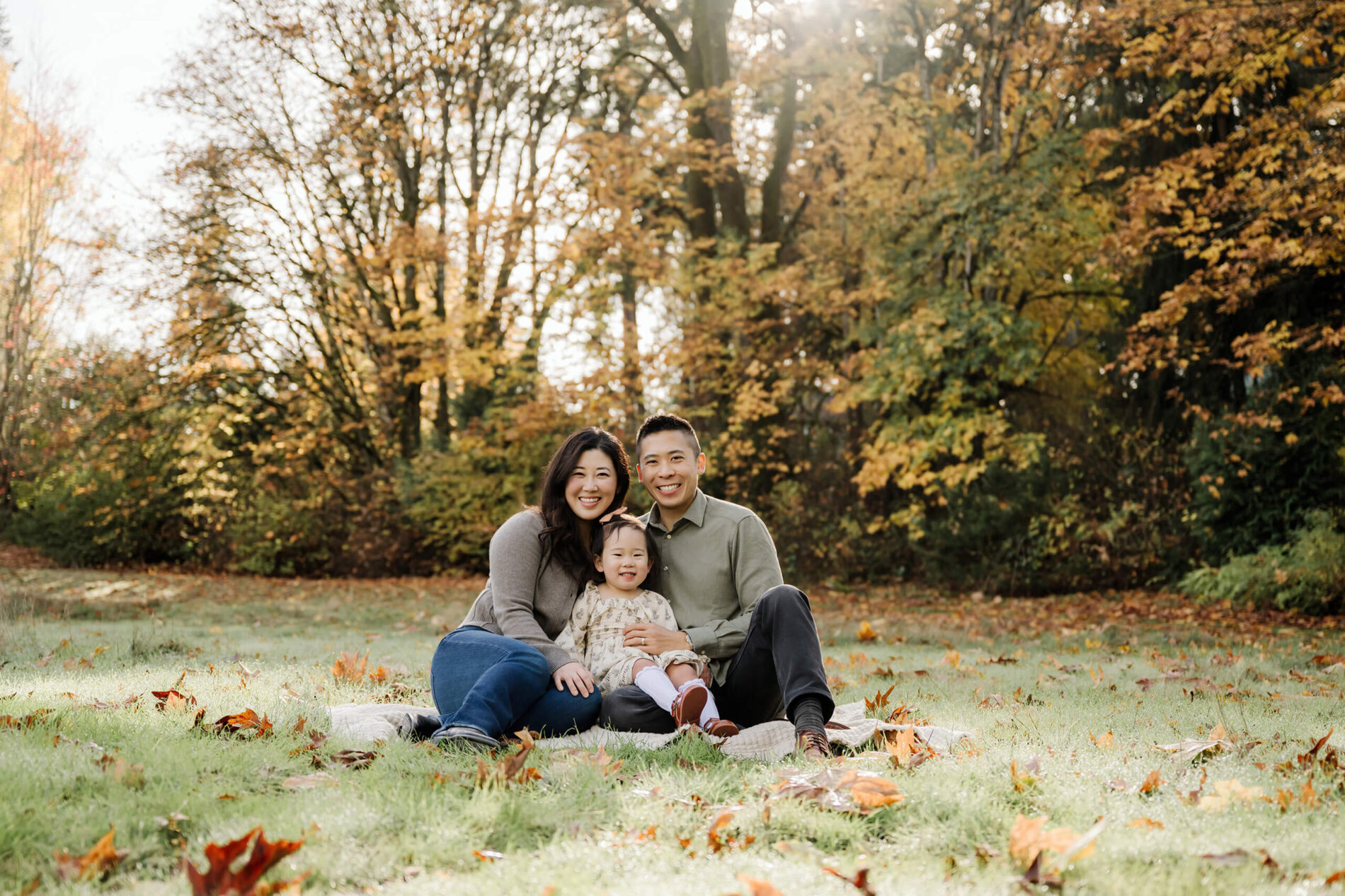 Family of three sits together on a blanket in a grassy clearing with fall trees behind them.