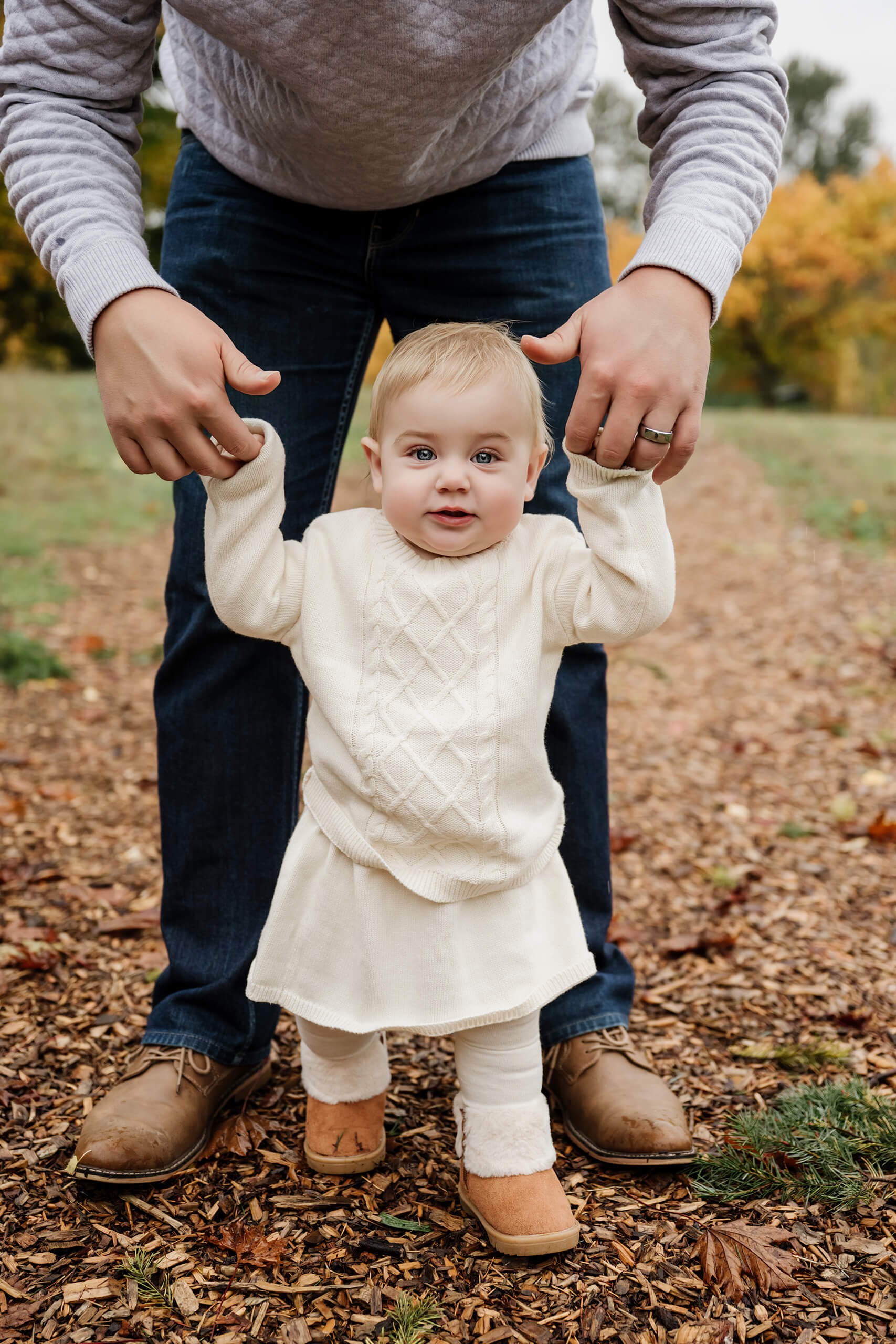 Baby takes steps while holding dad’s hands on a path surrounded by autumn color.