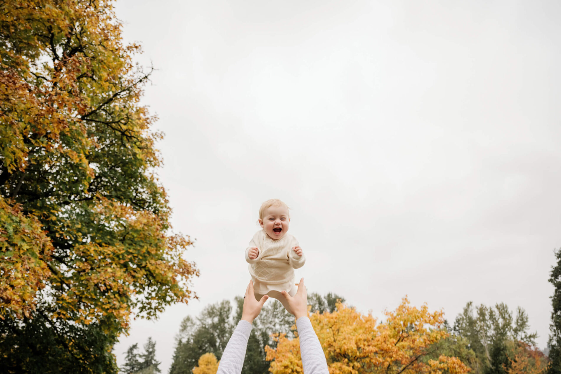 Laughing baby is lifted overhead against orange and yellow fall trees at Juanita Bay Park.