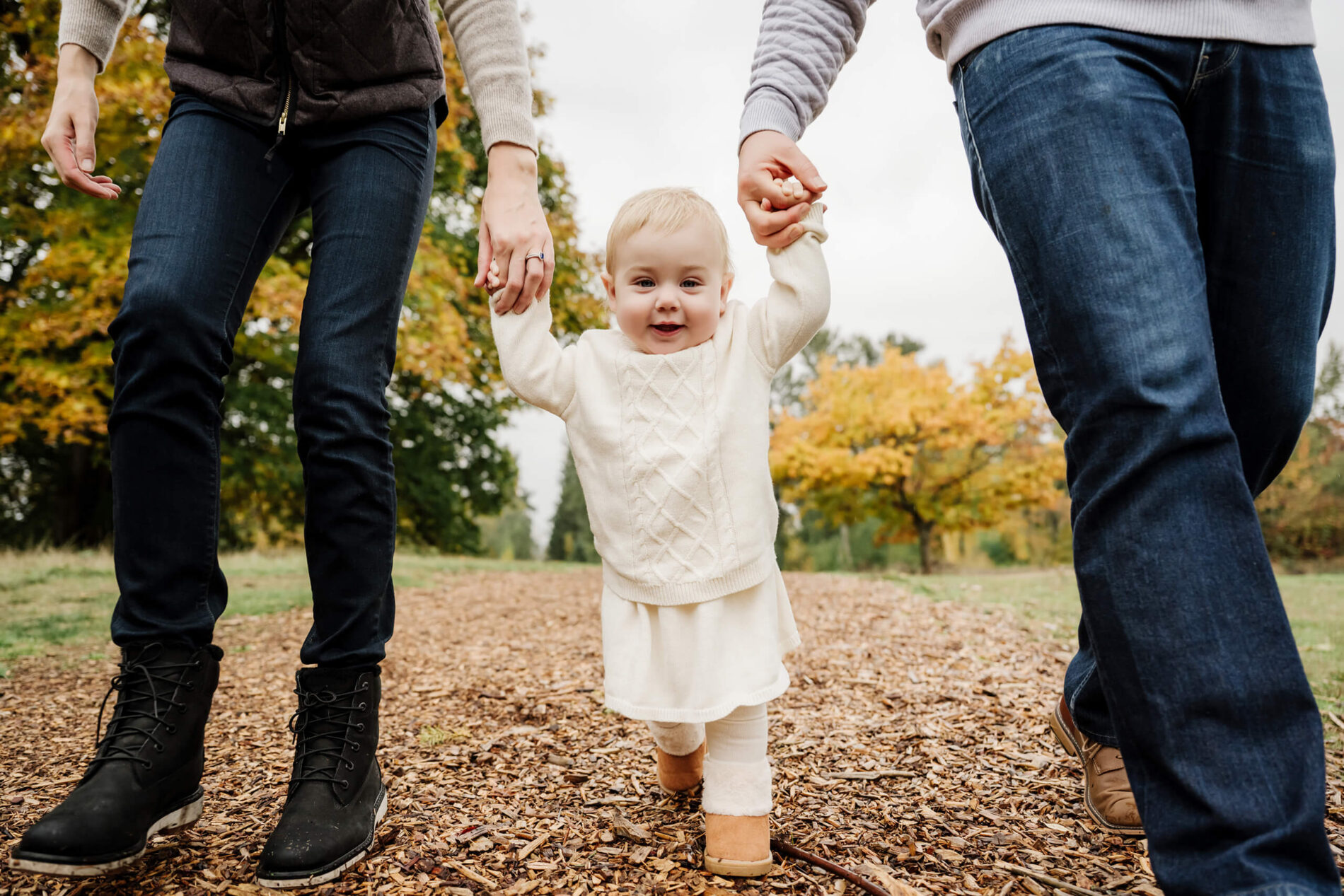 Baby walks toward the camera while holding both parents’ hands on a fall park path.