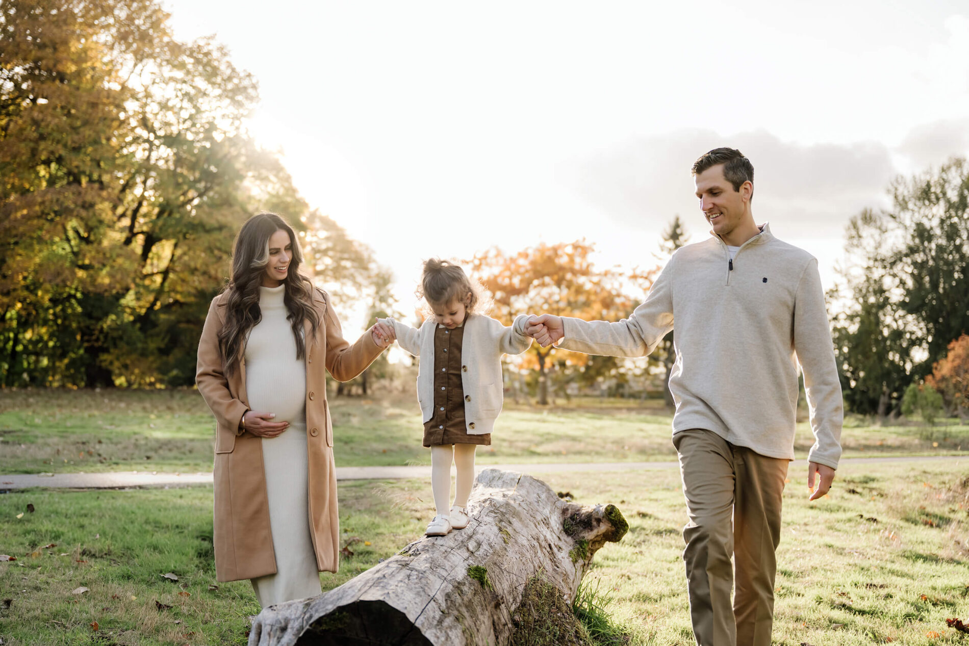 Parents hold their daughter’s hands as she balances along a fallen log in the autumn park.