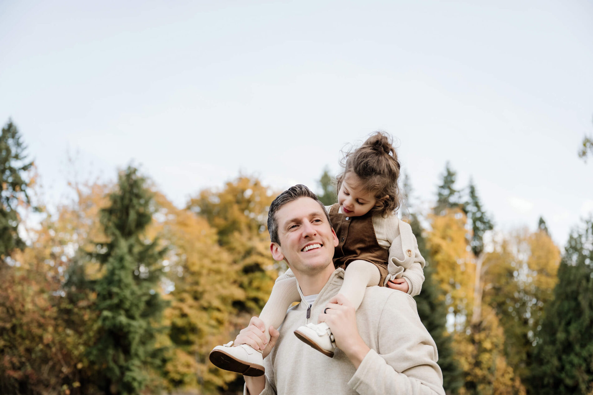 Dad carries his young daughter on his shoulders with golden autumn trees in the background.