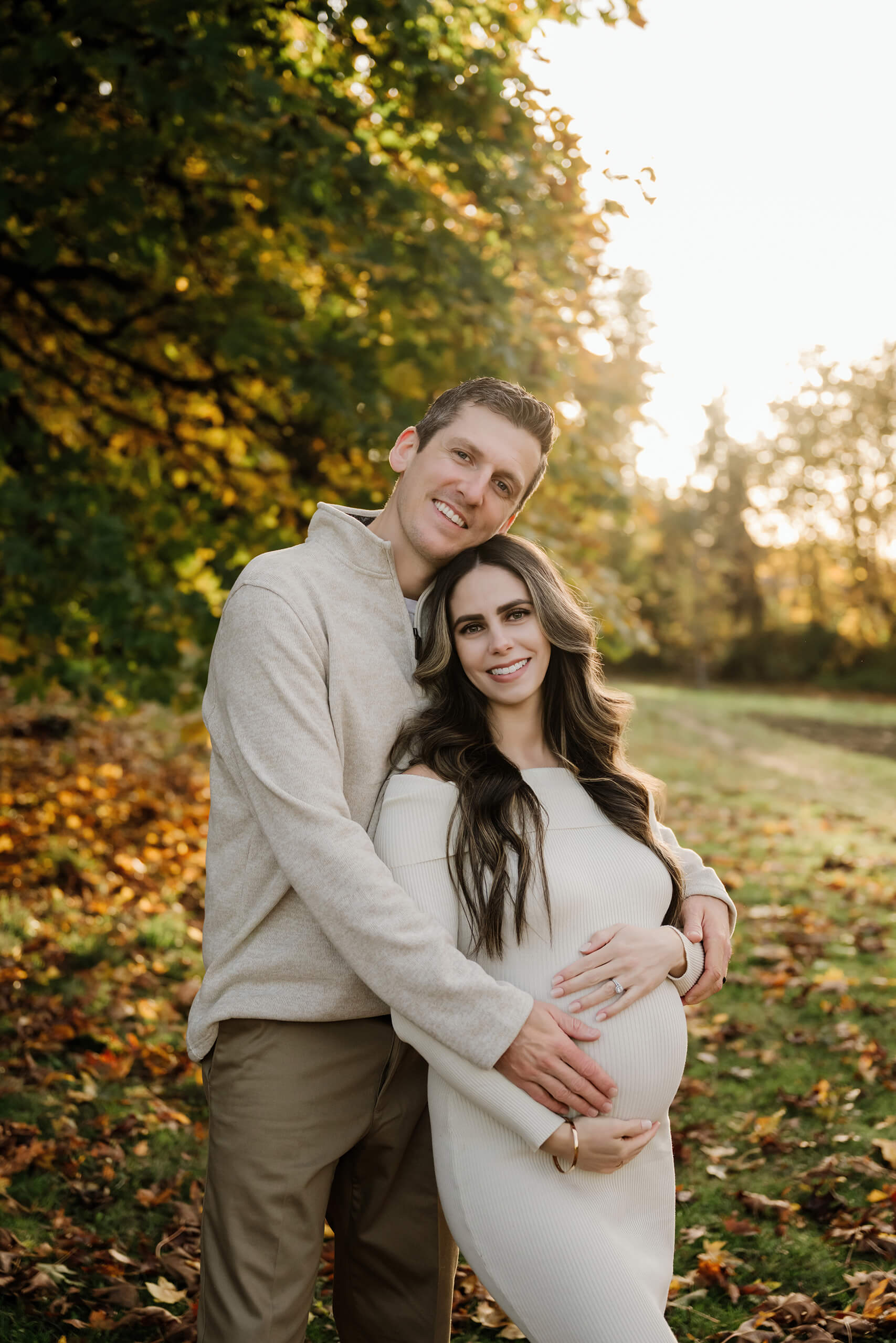 Expecting mom and her partner embrace beneath green and golden autumn leaves in warm evening light.