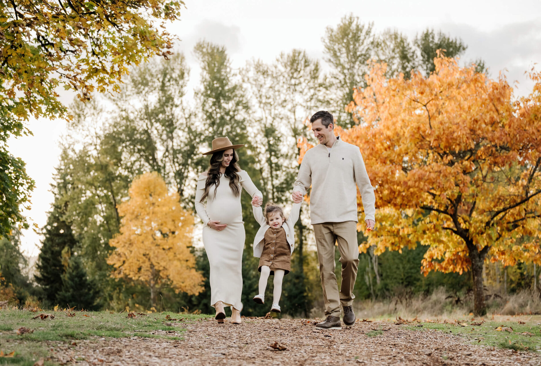 Expecting mom and her partner swing their daughter between them while walking through bright fall foliage.