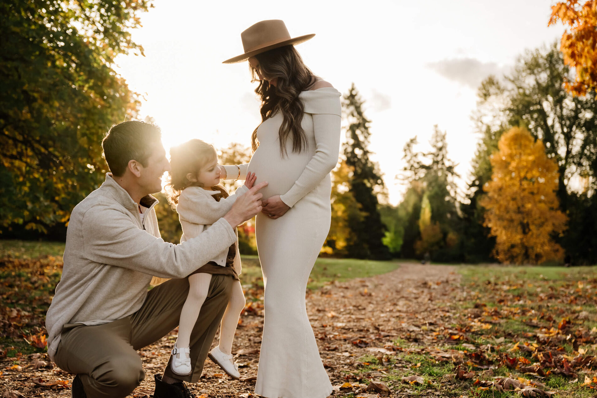 Expecting mom stands with her partner and daughter as they reach toward her belly in warm fall light.