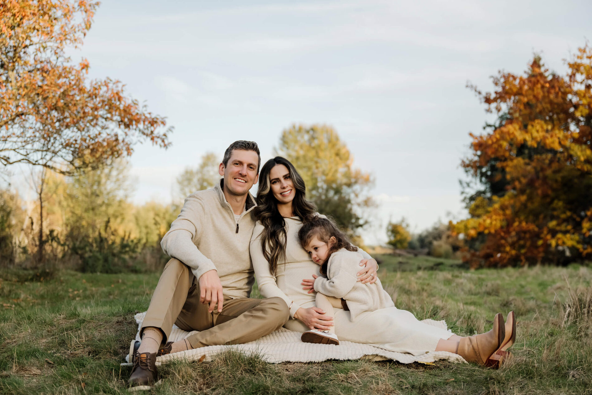 Expecting mom sits with her partner and daughter on a blanket surrounded by autumn trees.