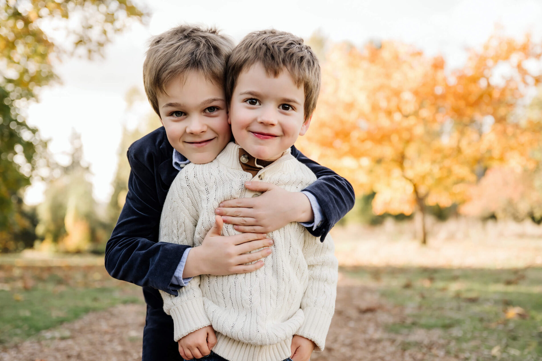 Two brothers hug closely with bright orange fall foliage behind them.