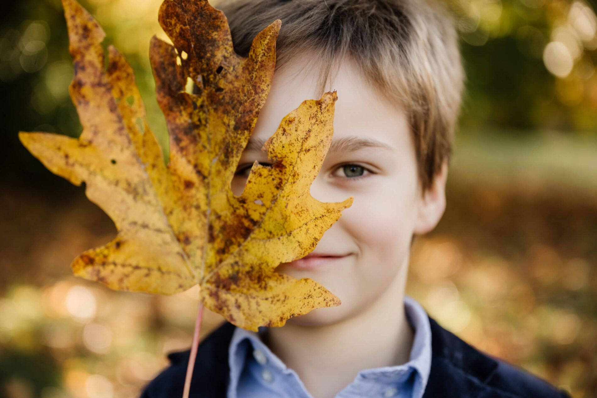 Young boy holds a large yellow fall leaf in front of one eye.