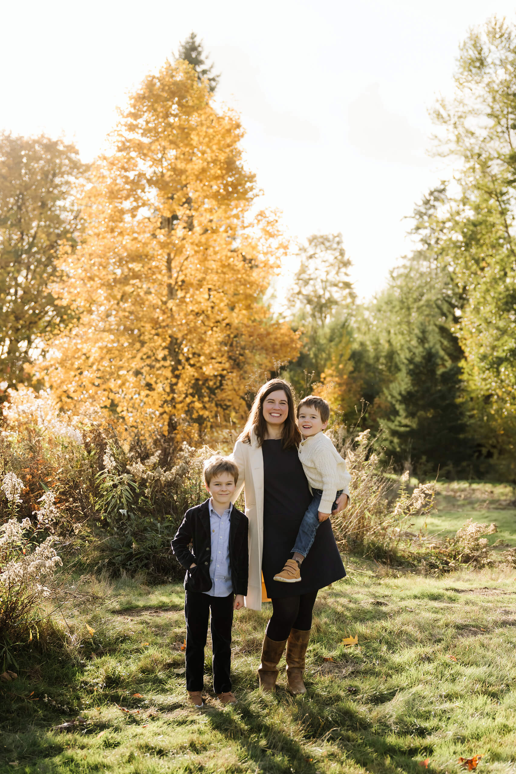 Mom stands with her two sons beside tall grasses and a glowing yellow autumn tree.