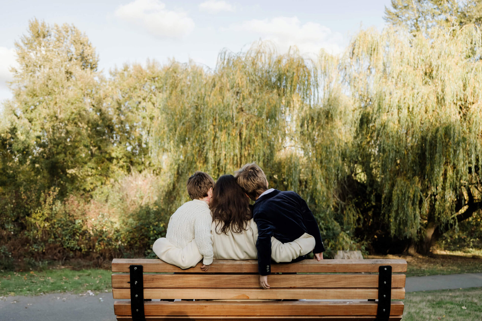 Mom sits between her two sons on a park bench beneath tall willow trees.