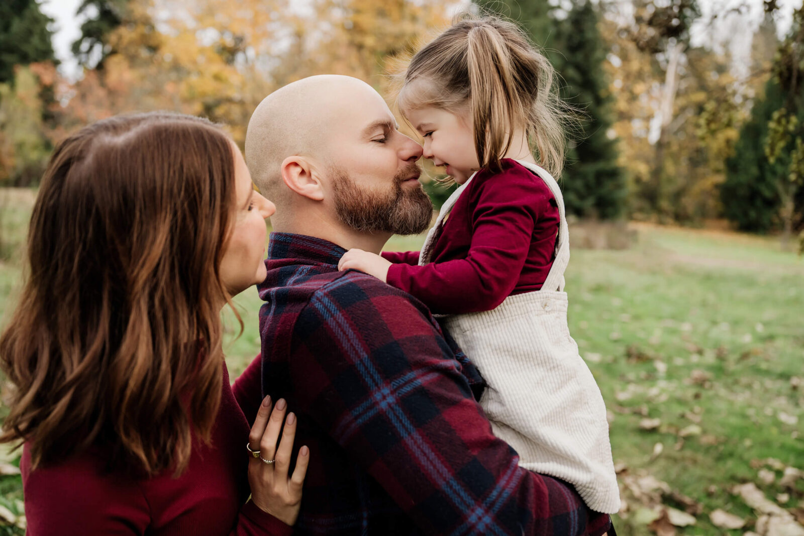 Dad holds his young daughter close as they touch noses, with mom beside them in an autumn park.