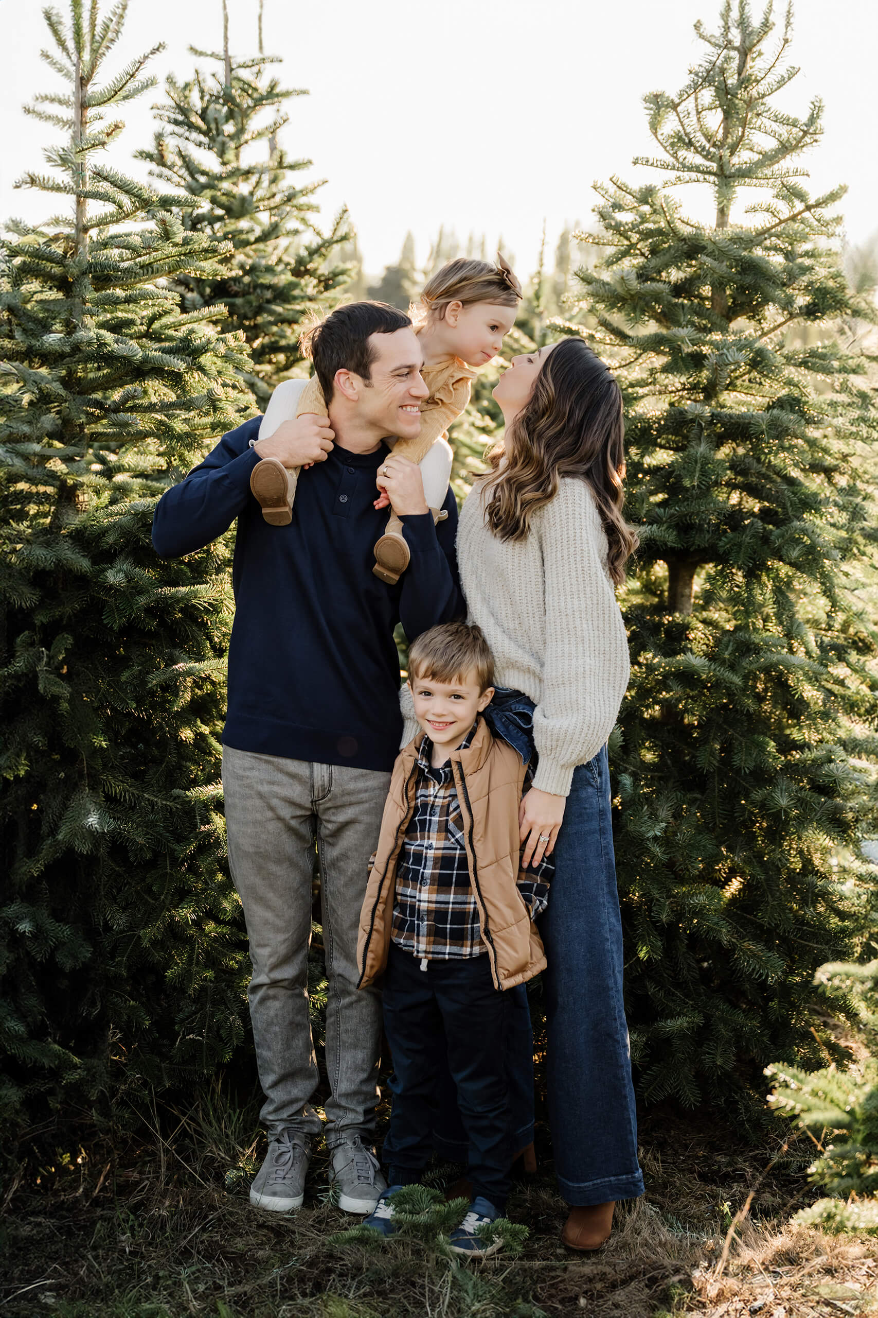 Mom and dad stand with their son and daughter among evergreen trees as their daughter sits on dad’s shoulders and leans toward mom.