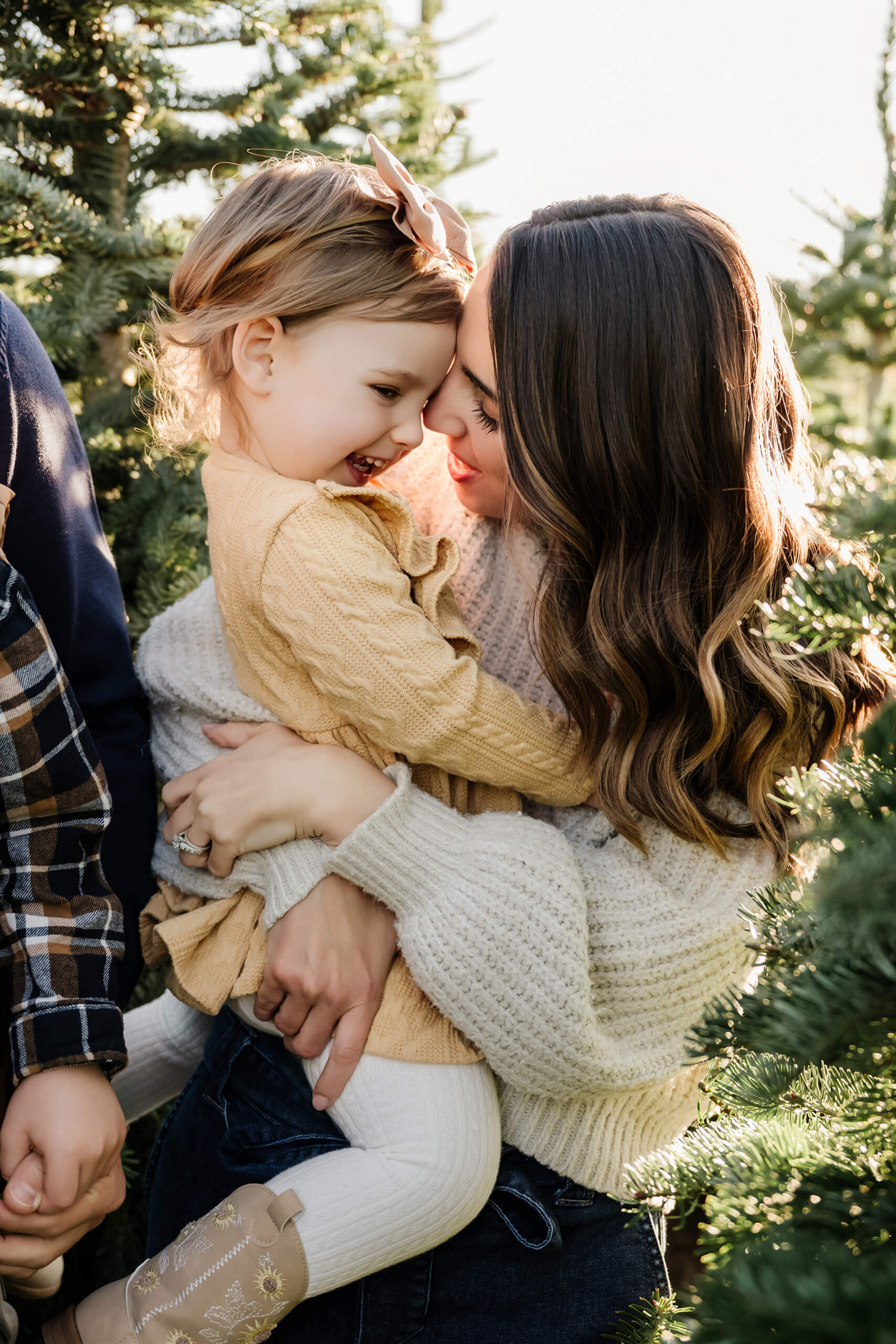 A mom holds her daughter close as they touch foreheads and smile among evergreen branches.