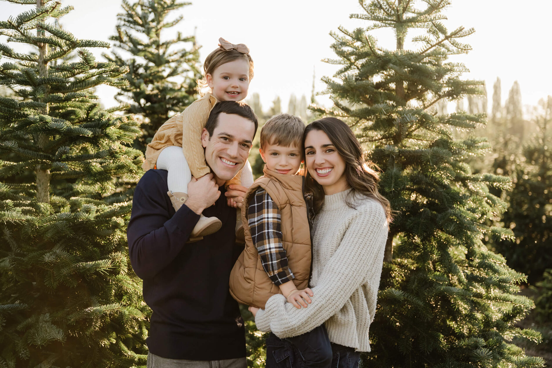 Mom and dad pose with their son and daughter between evergreen trees, with their daughter sitting on dad’s shoulders.