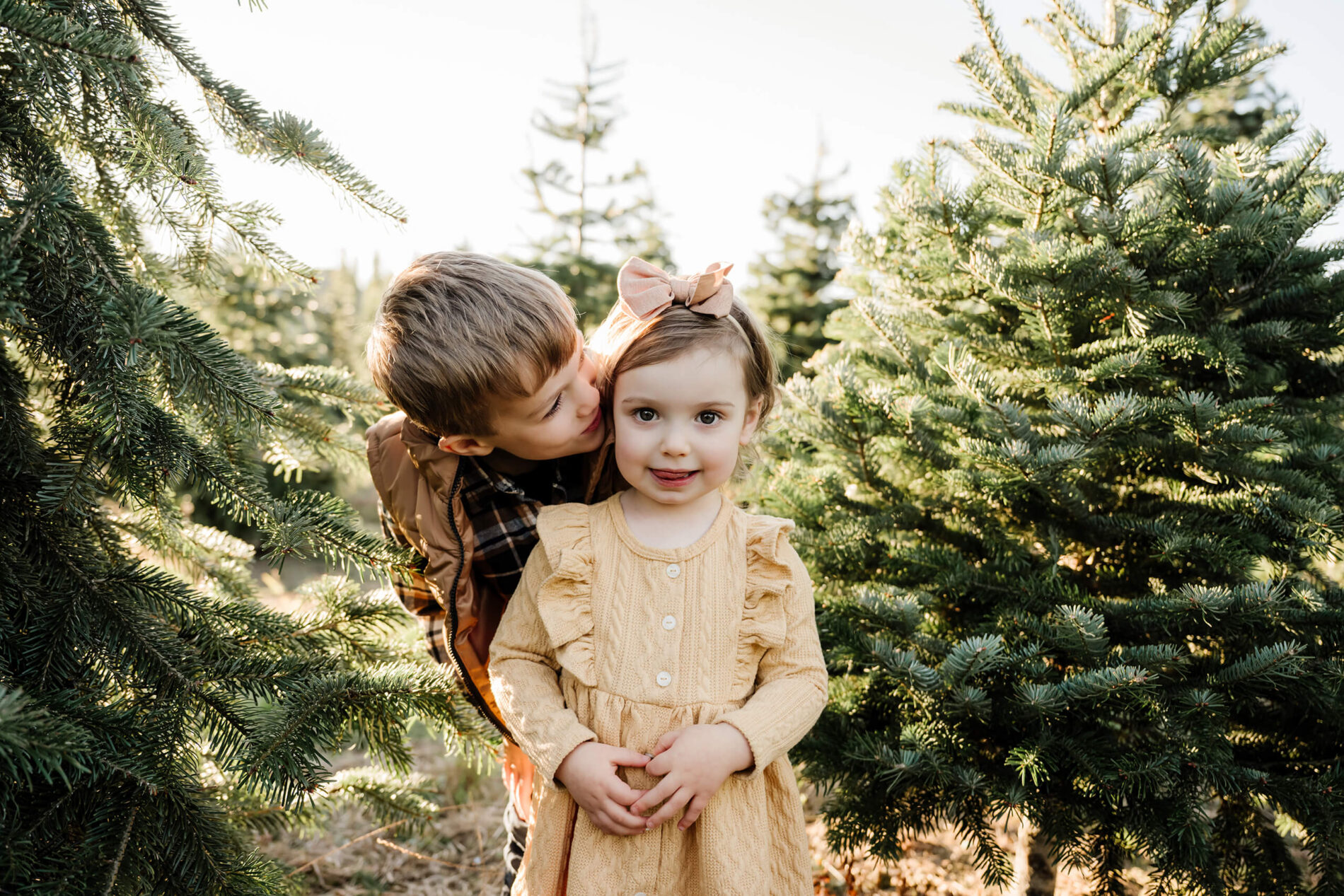 A brother leans in toward his sister as she looks at the camera between evergreen trees.