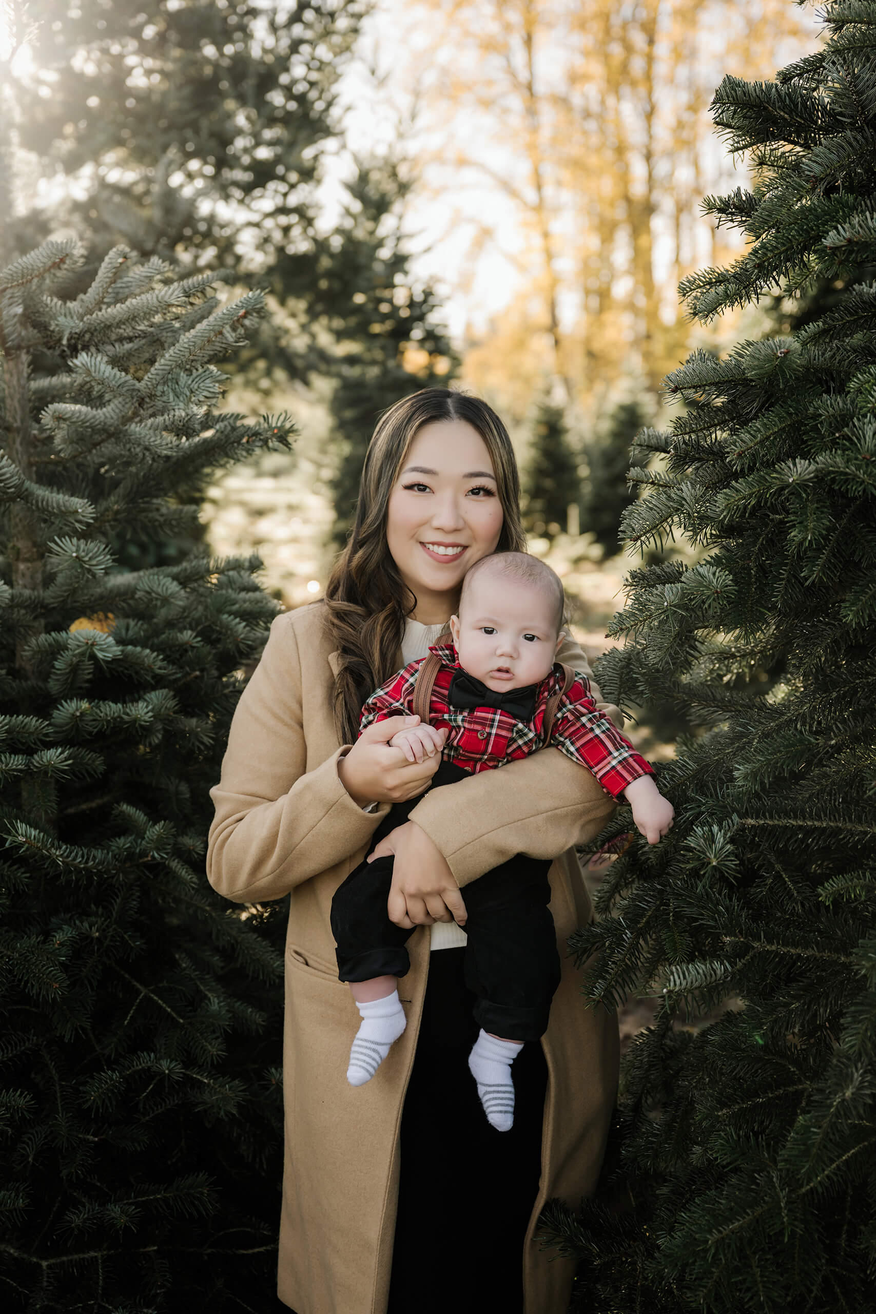 A mom holds her baby between evergreen trees, both facing the camera with golden foliage in the background.